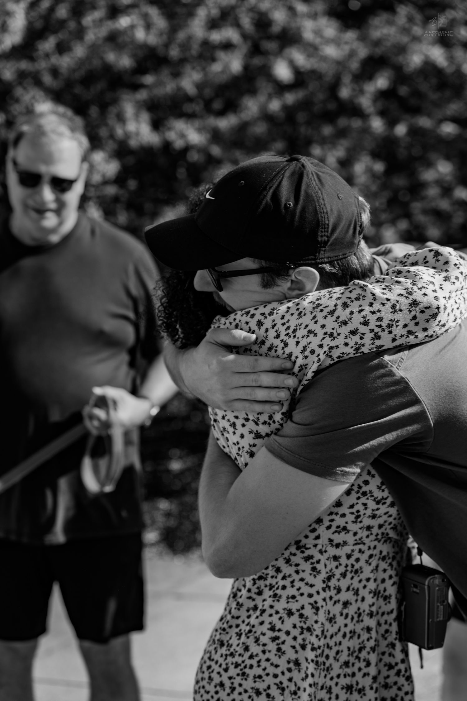 Two people share a heartfelt embrace outdoors while a man watches in black and white.