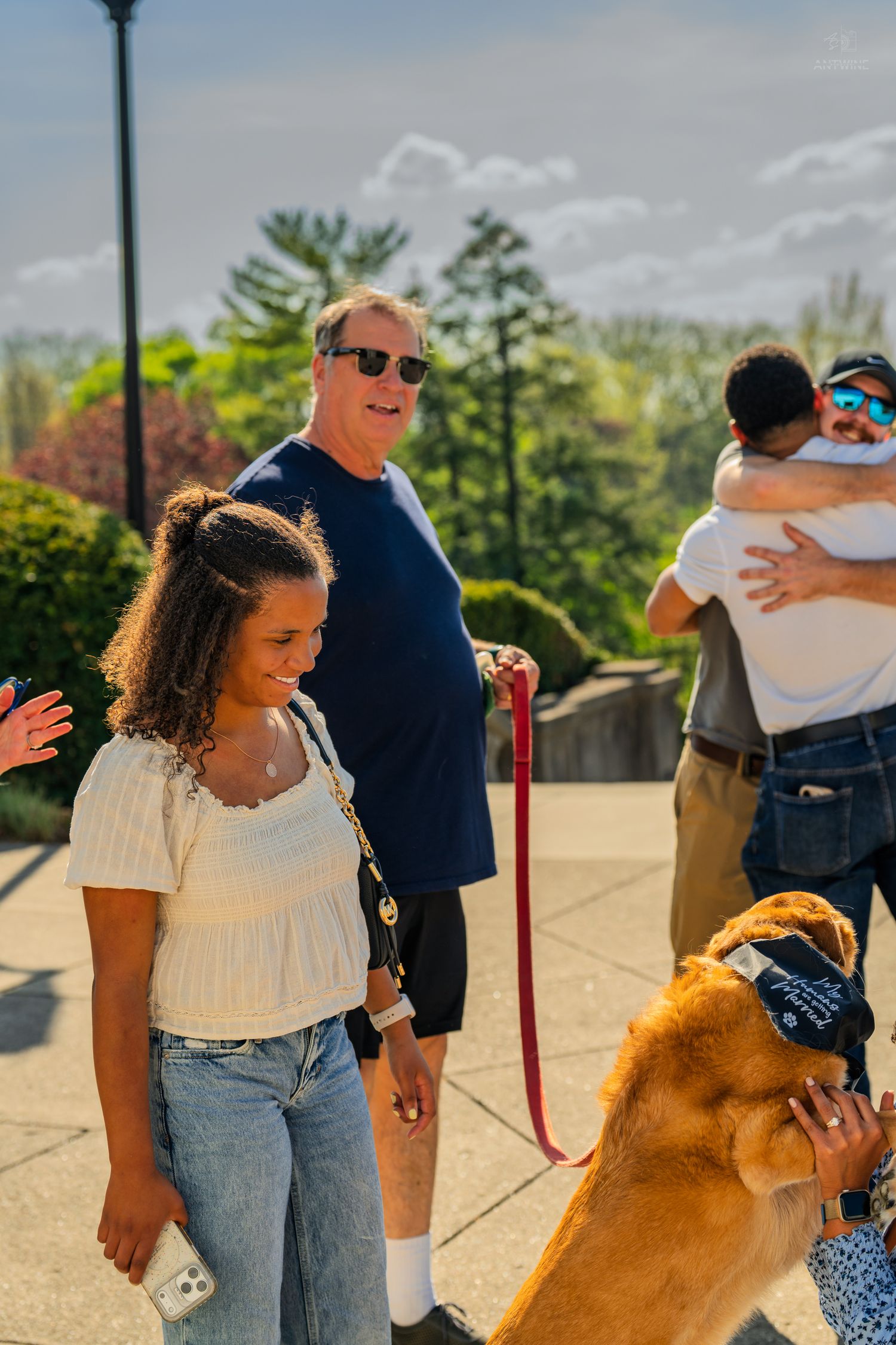 A smiling young woman in a white top stands near an older man holding a dog on a red leash outdoors.
