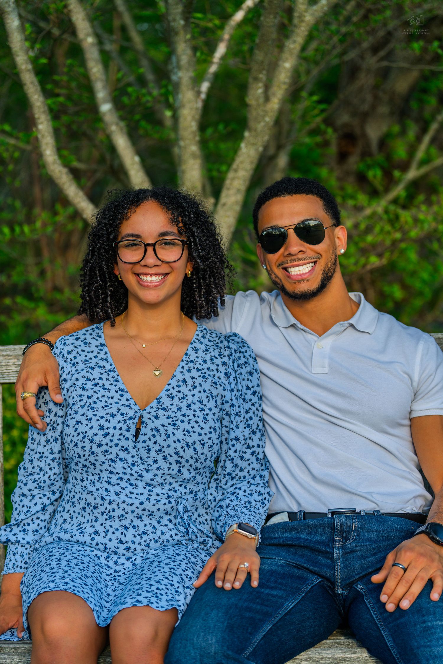 Happy couple sits on a bench outdoors, woman in blue floral dress and man in white polo shirt.