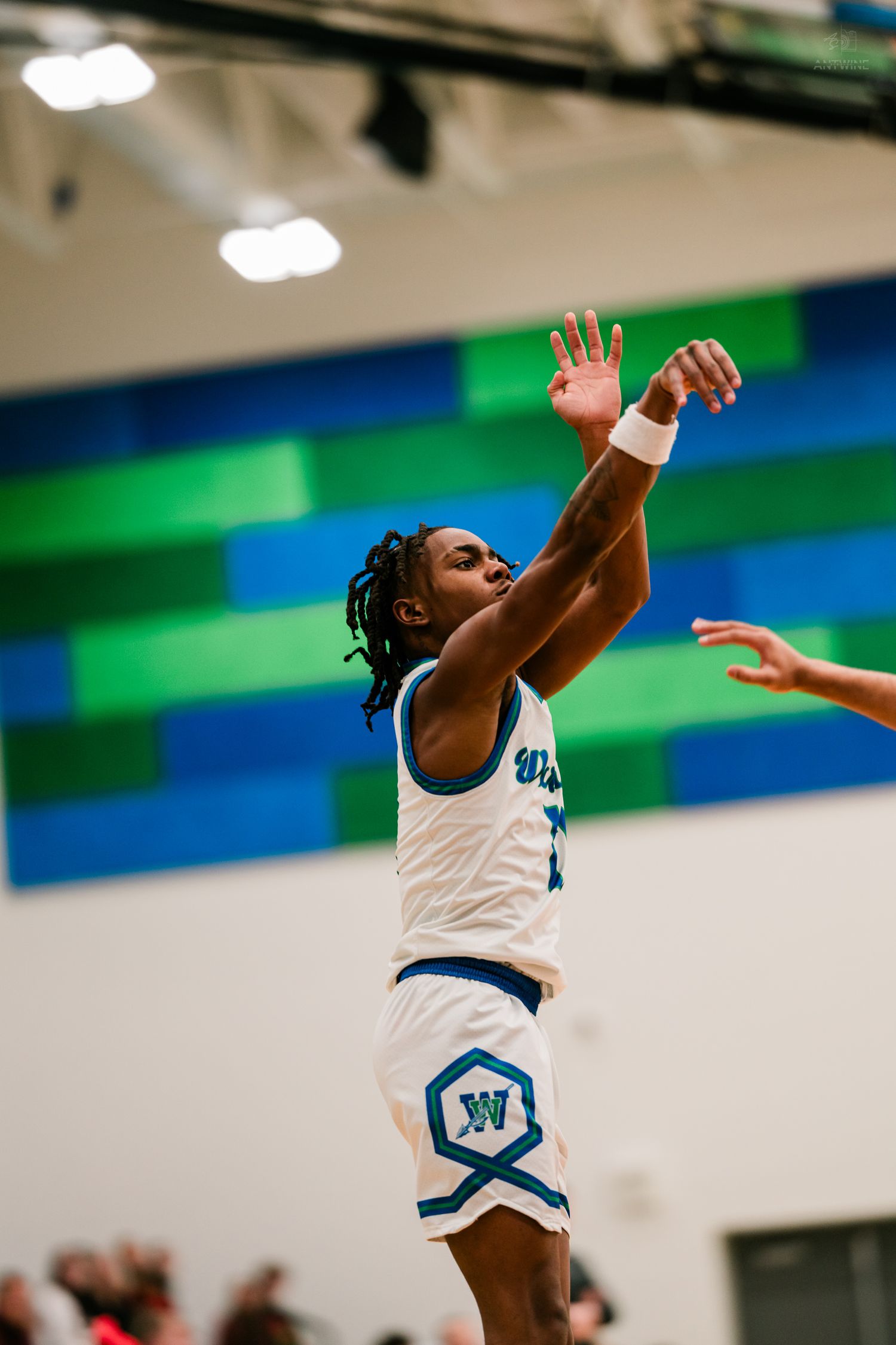 A Warriors basketball player jumps to shoot during a high school game in a gym with blue and green wall art.