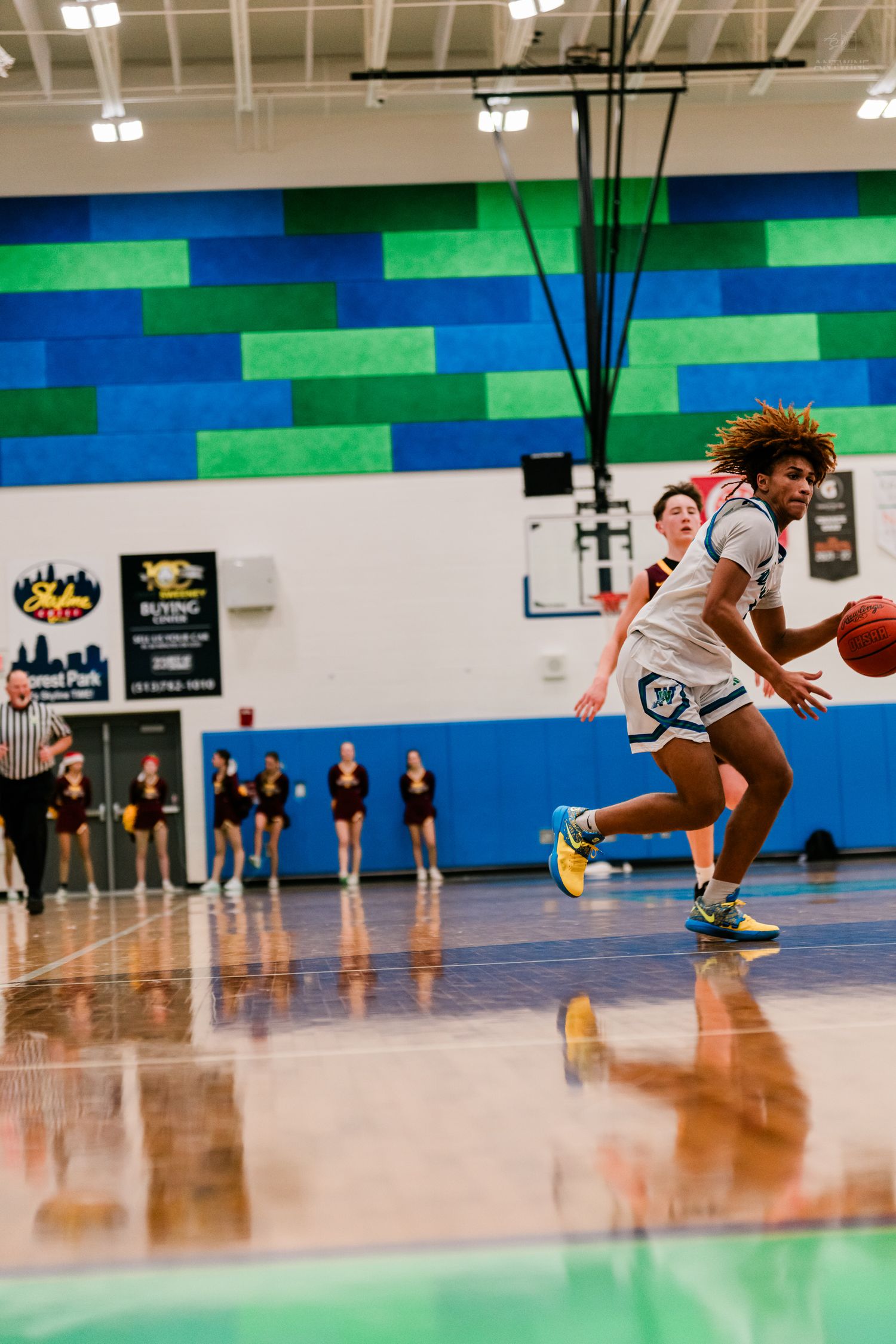 A Warriors basketball player dribbles down the court during a high school game in a blue and green gymnasium.
