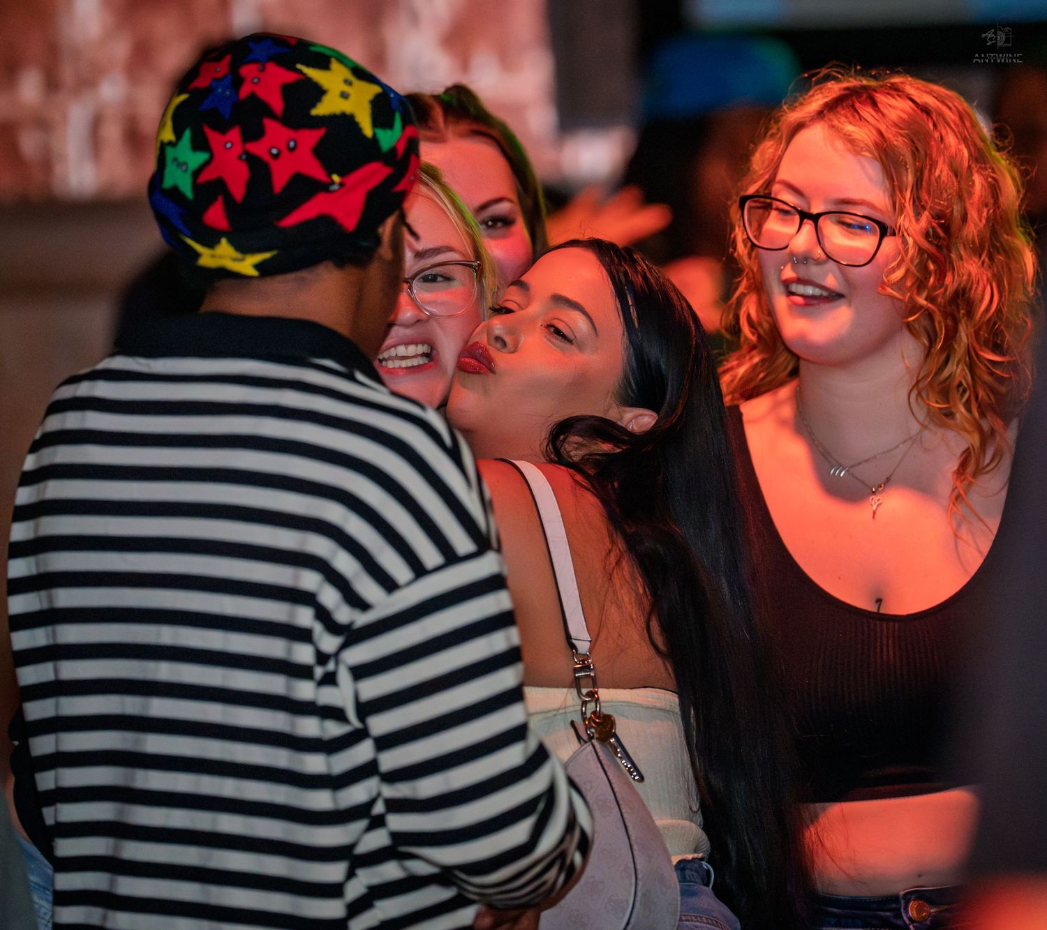 Young women smiling and socializing with a man in a colorful star-print hat at a club.