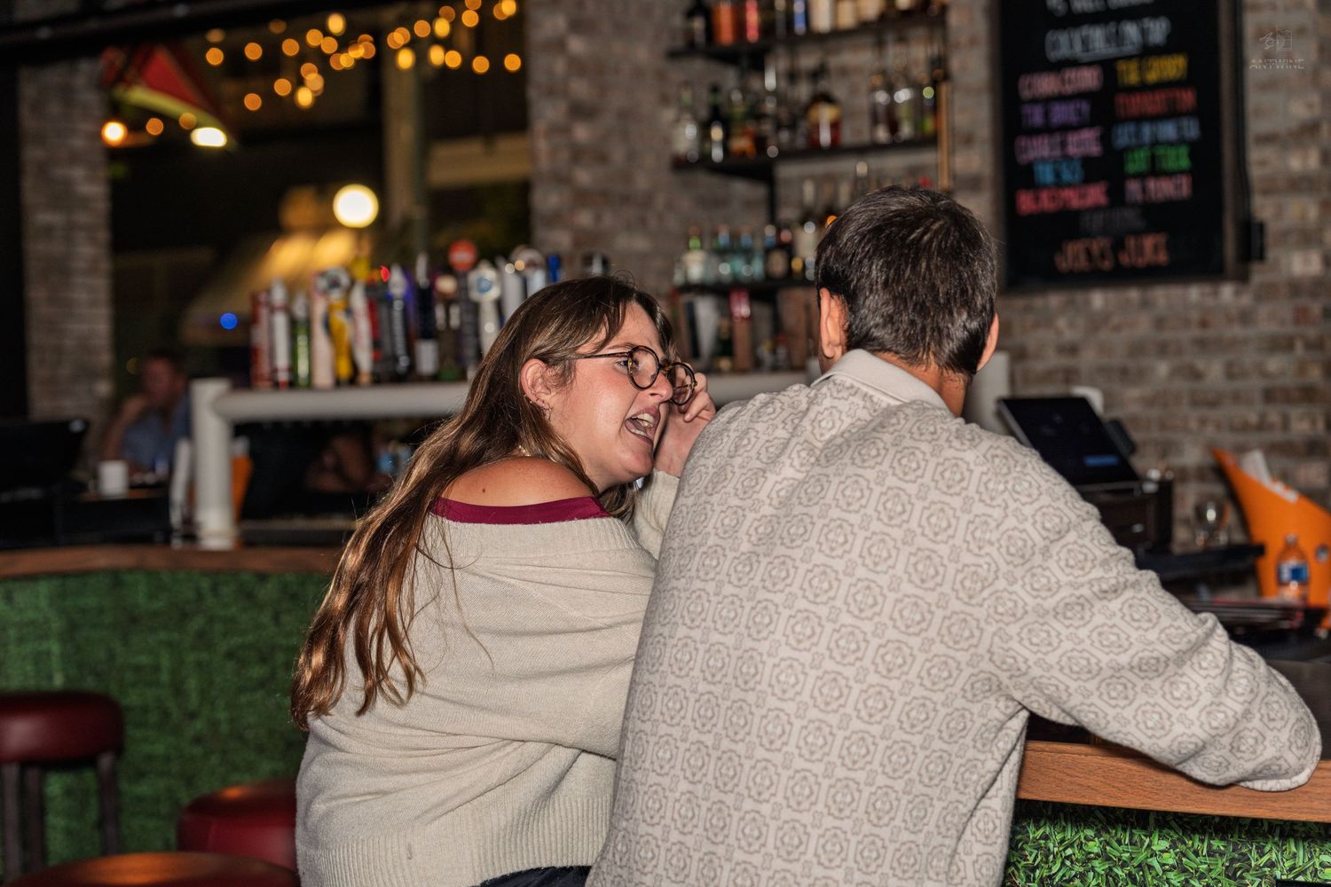 Couple sharing a private conversation while leaning against a bar with string lights overhead.