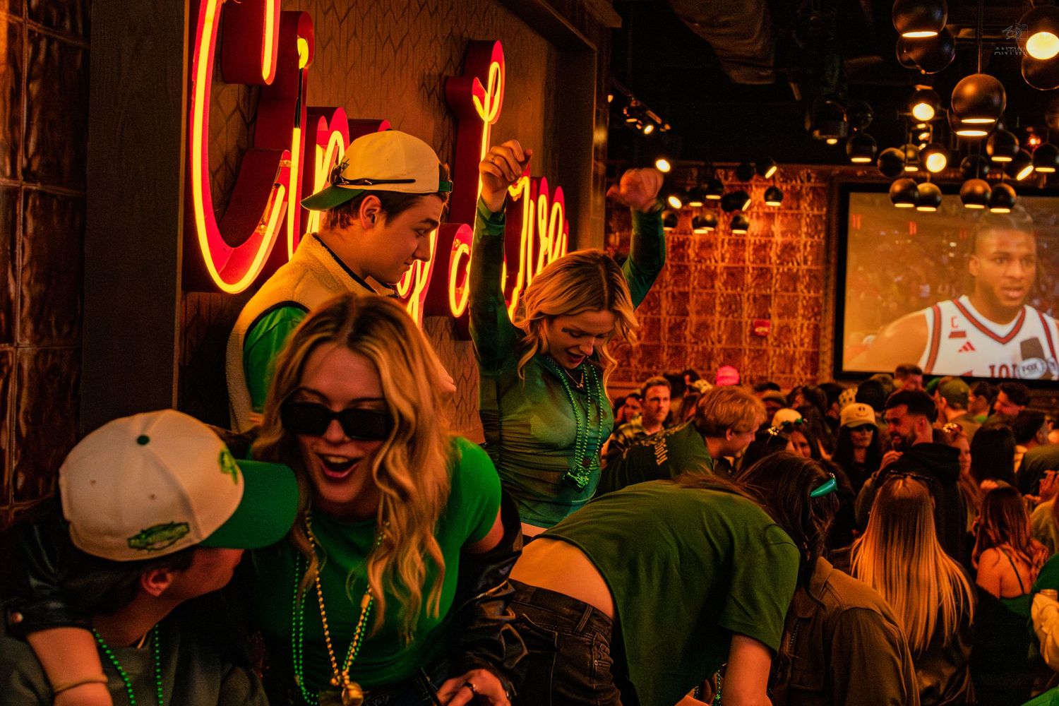 Crowd of people wearing green celebrating St. Patrick's Day at a lively bar with neon signs.