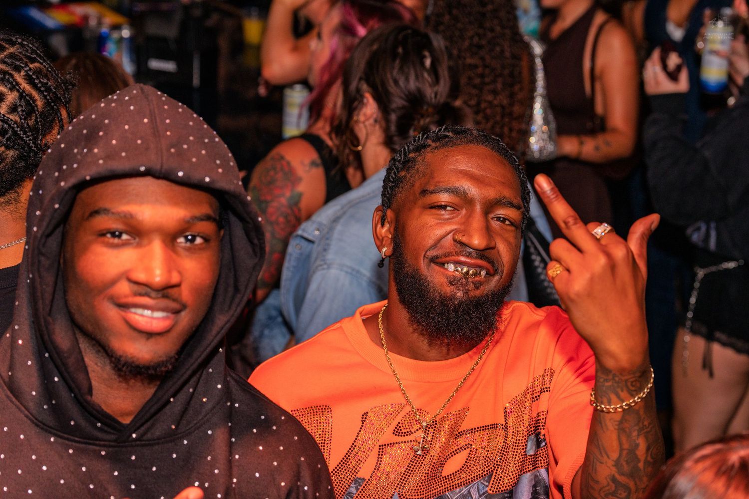 Two smiling men posing at a lively party, one flashing a hand sign in orange shirt.