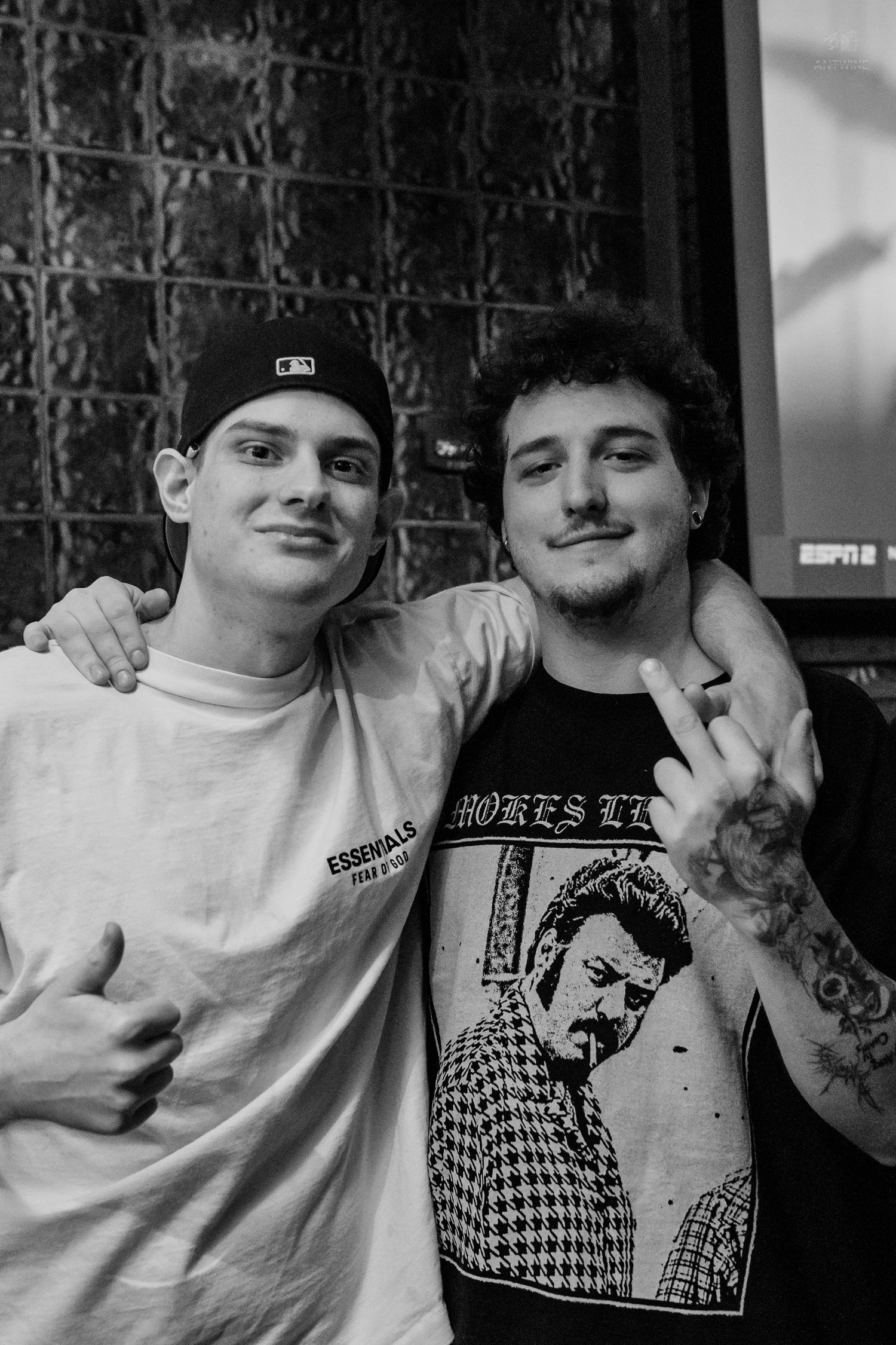 Black and white photo of two young men posing together, one flashing a peace sign at a bar.