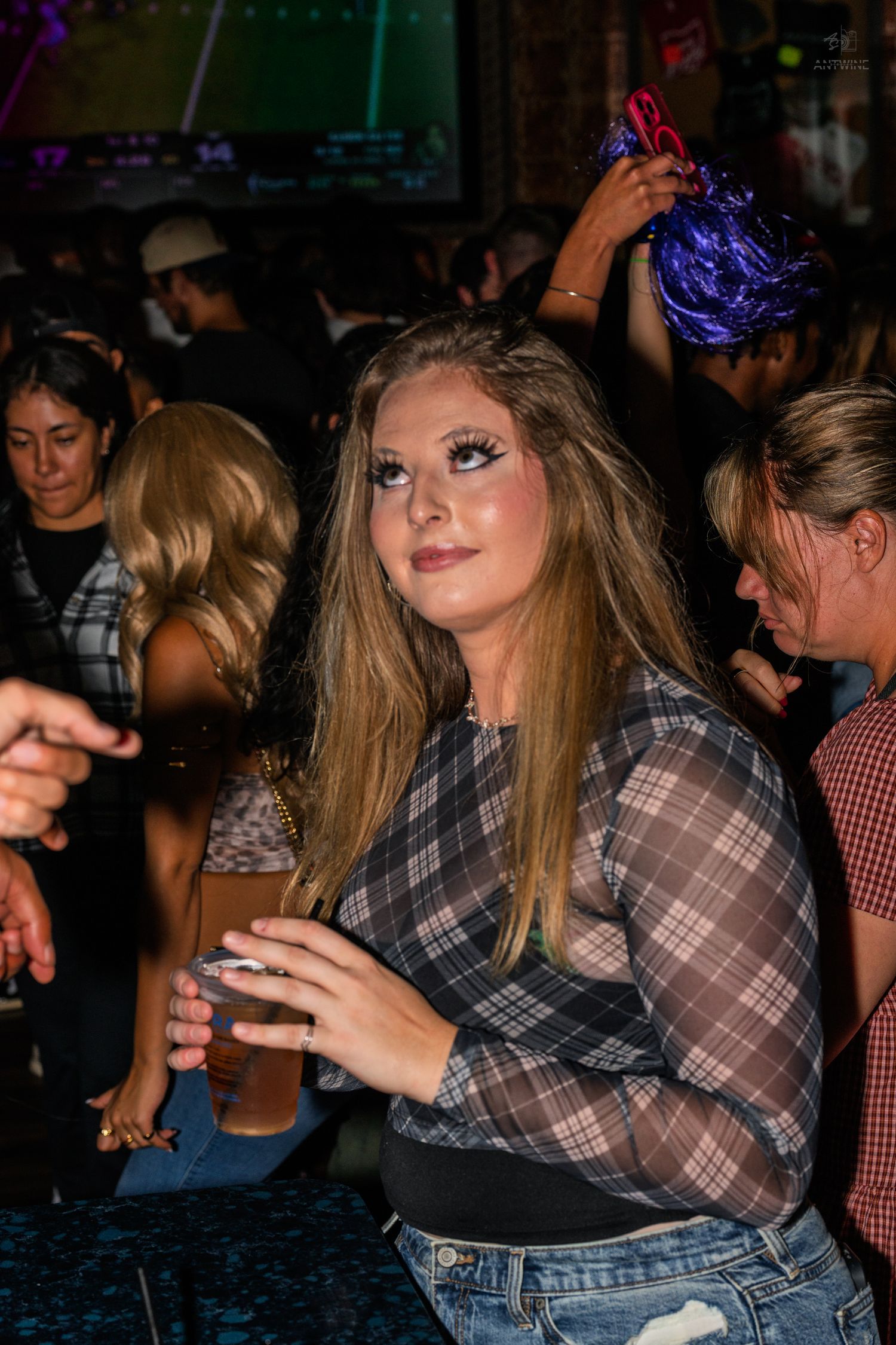 Young woman in plaid shirt holding drink at crowded nightclub party.