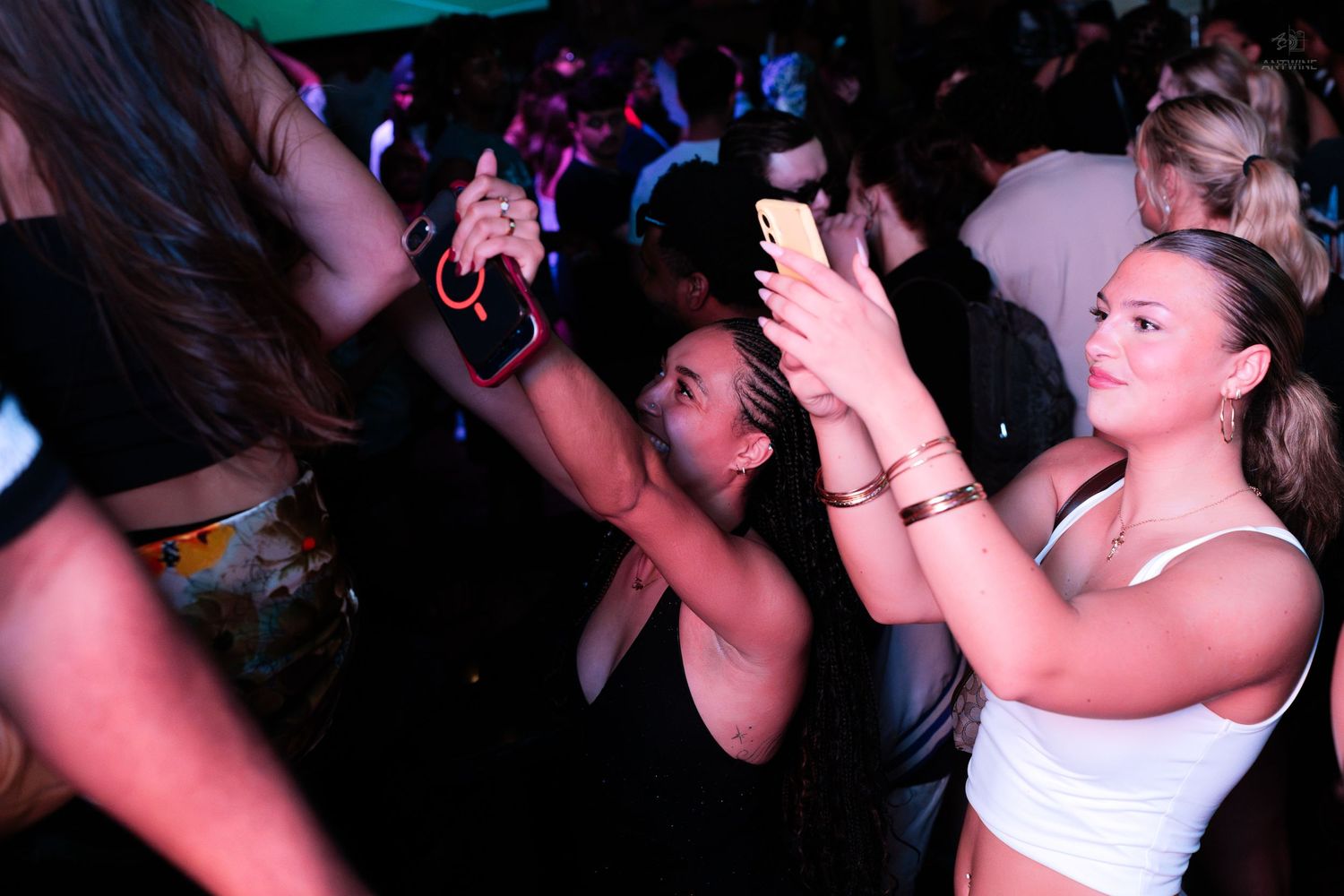 Two young women taking photos with smartphones on a packed nightclub dance floor.