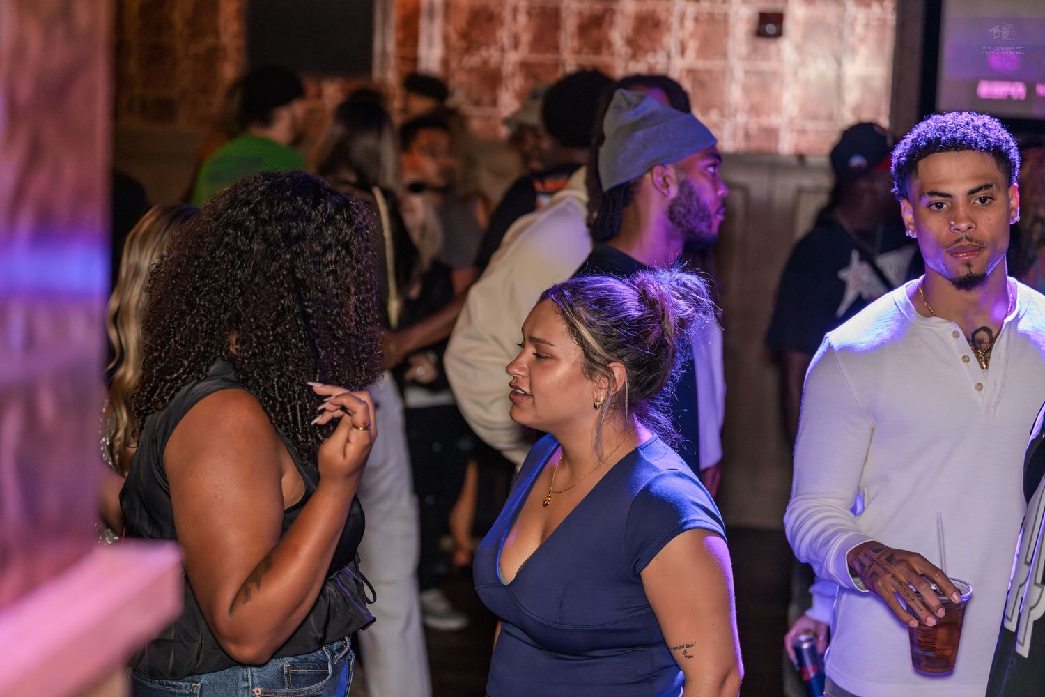 Young adults mingling at a dimly lit nightclub with purple lighting, one man holding a drink.