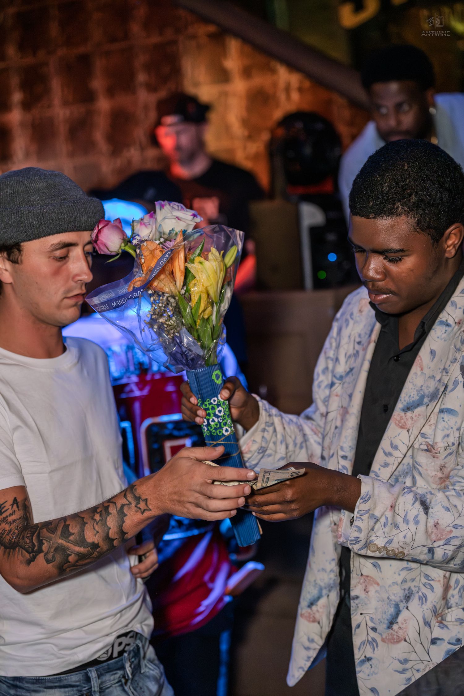 Two men exchange flowers and a phone at a lively nightclub event with colorful lights.
