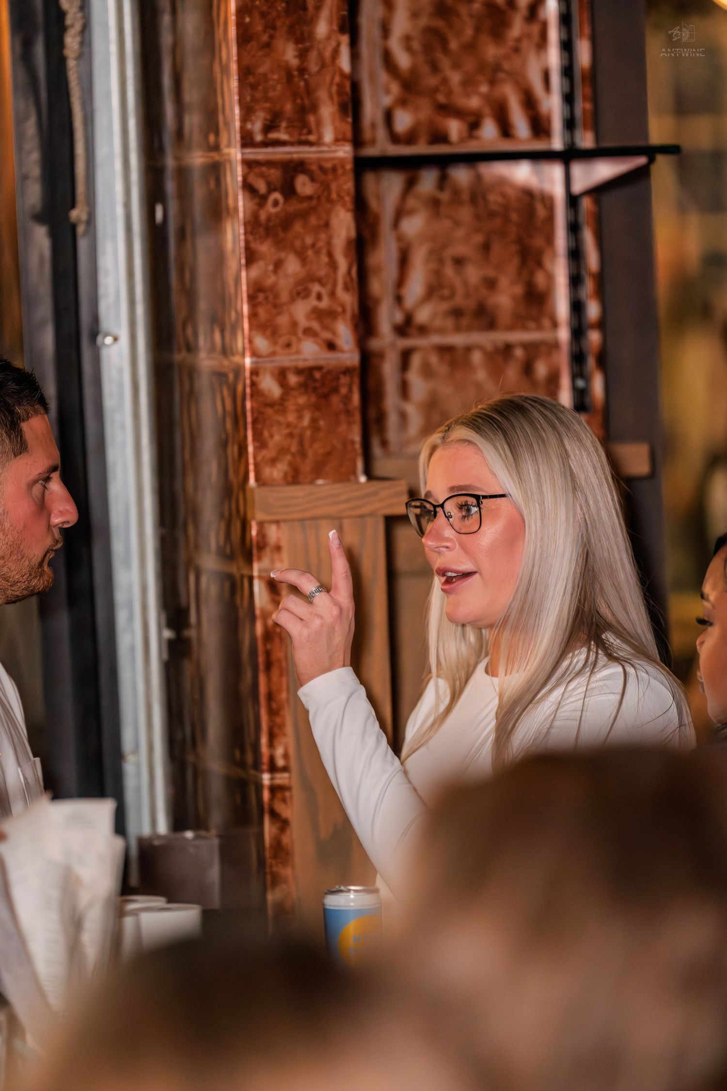 Blonde woman with glasses gesturing while talking at a restaurant bar setting.