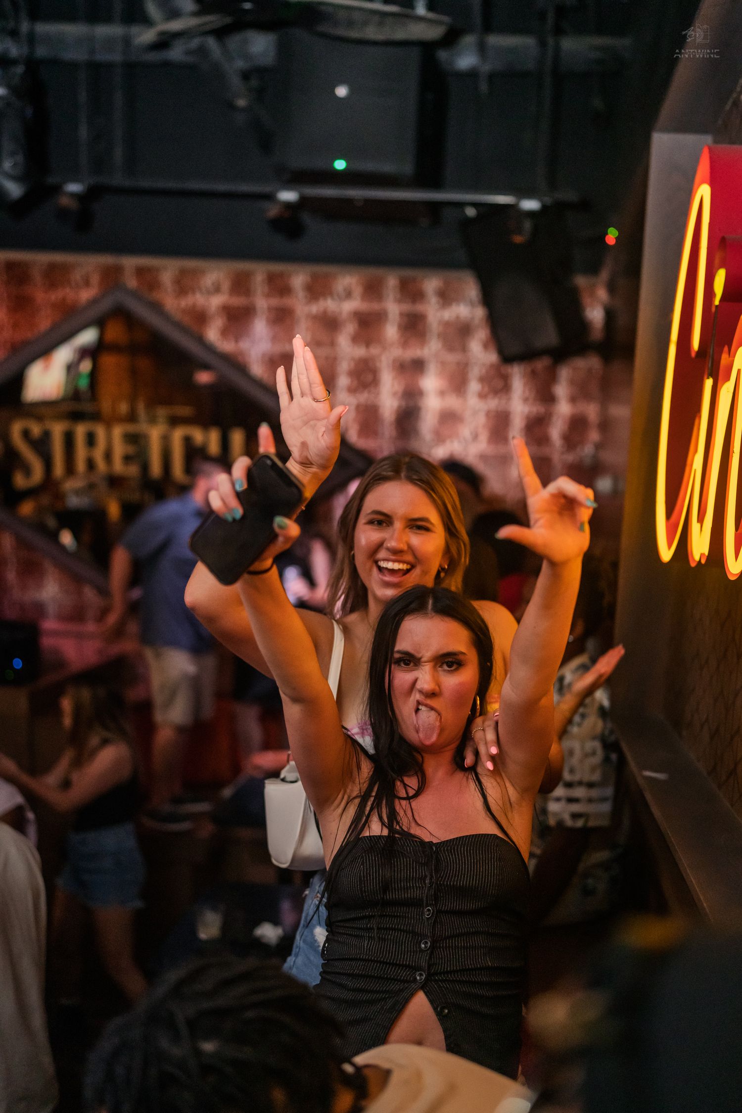 Two women posing energetically with hands raised near a neon sign at a nightclub.