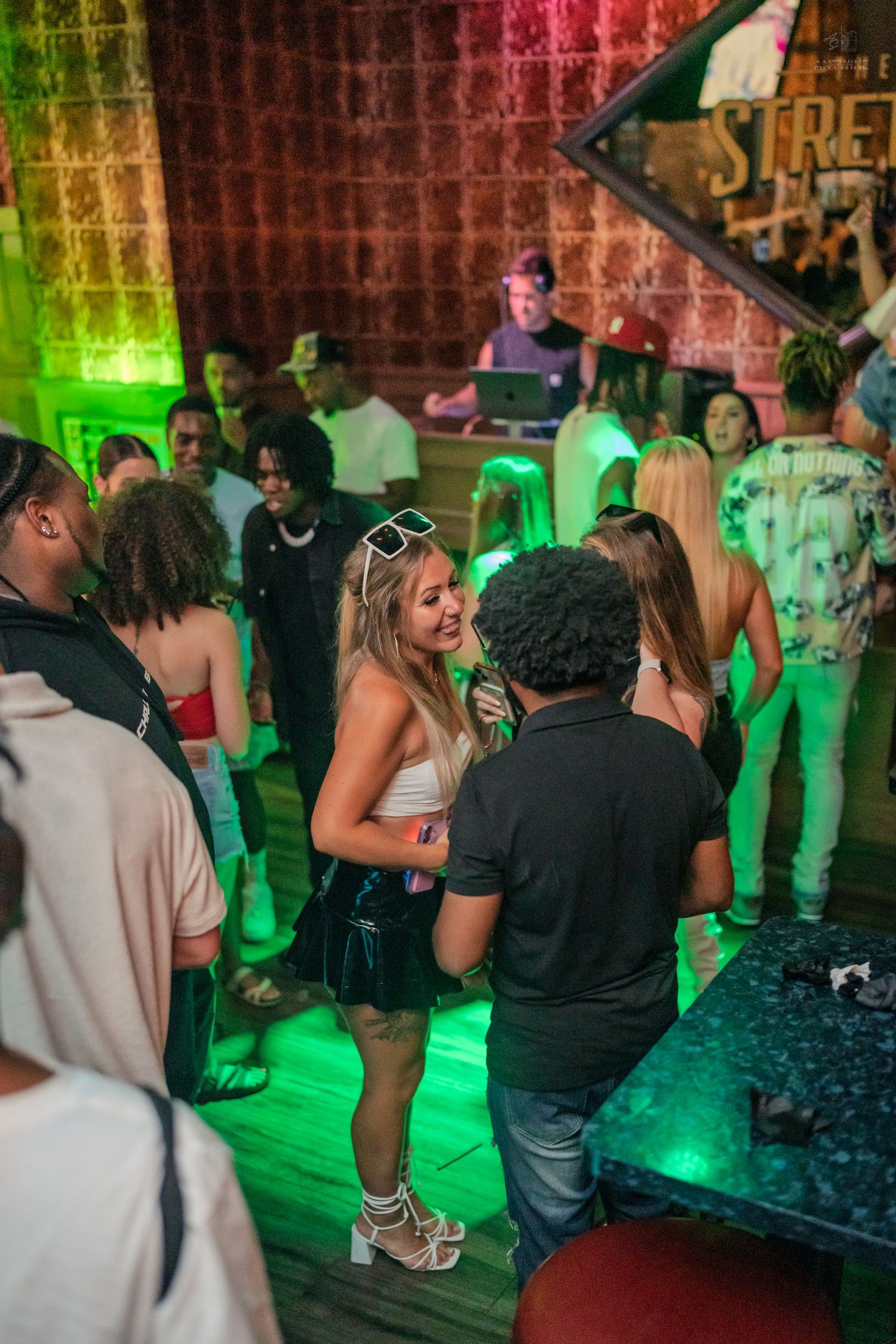 Smiling blonde woman in a mini skirt talking to someone near a DJ booth in green light.