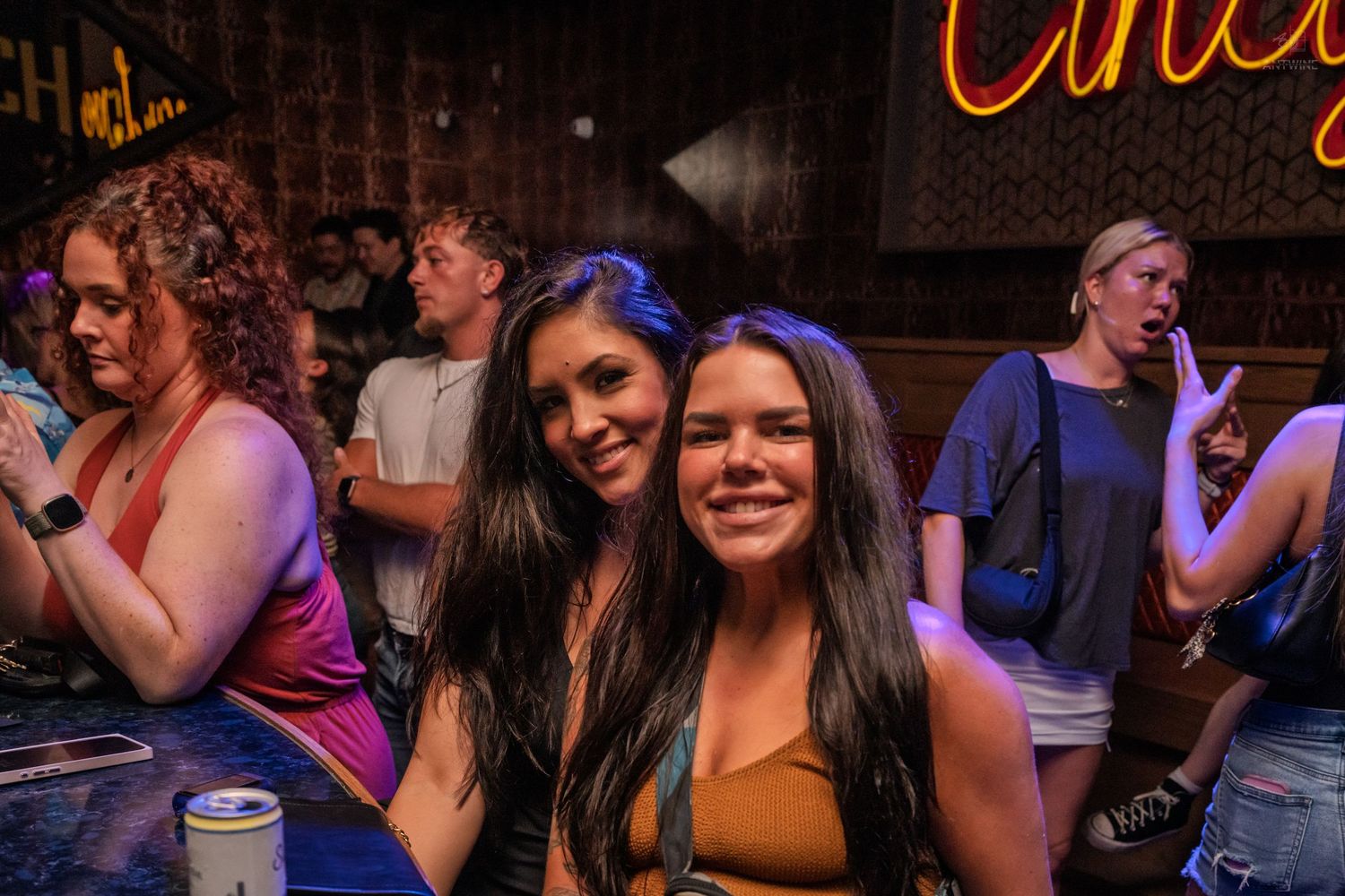 Two smiling women posing at a busy bar with colorful neon lighting.