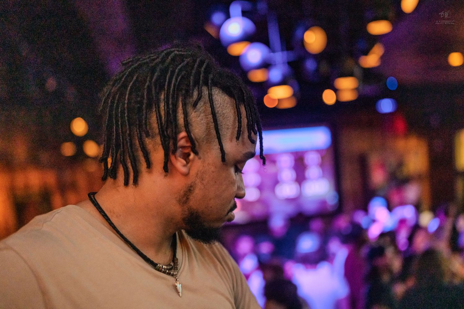 A man with dreadlocks bows his head in a purple-lit nightclub with blurred crowd in background.