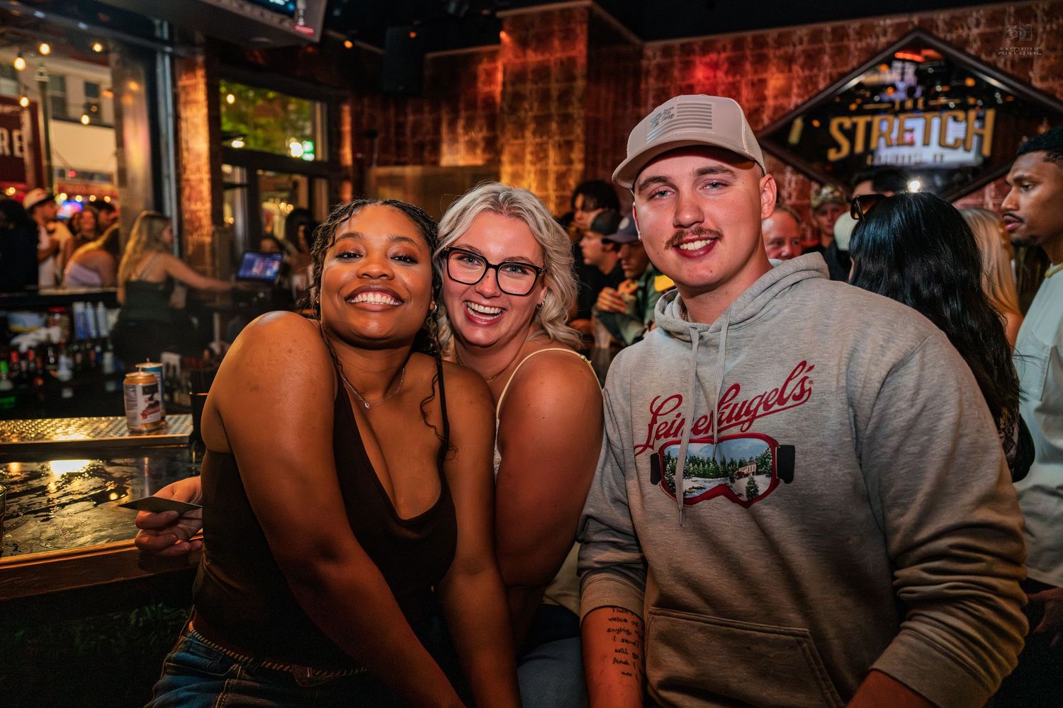 Three friends smiling and posing together at a busy bar under a Stretch sign.