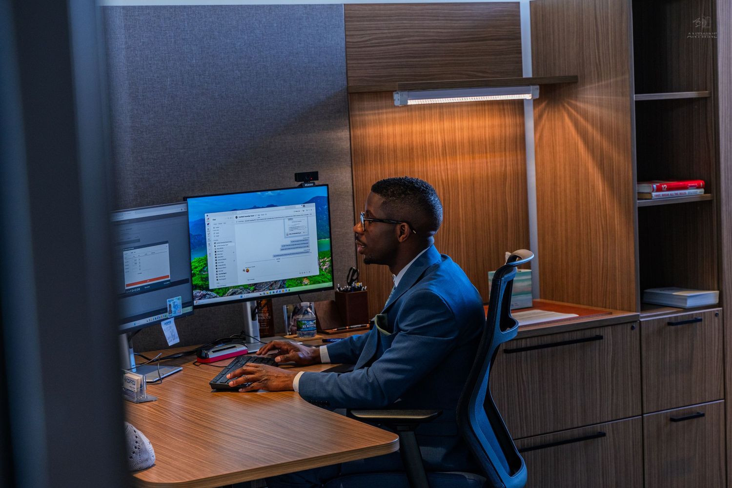 Professional Black businessman working at computer desk in modern office cubicle under warm lamp light.