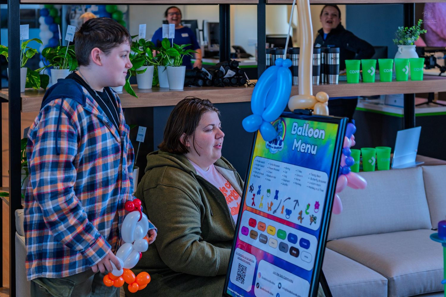 Two people browse a colorful balloon menu display at an indoor event with decorative plants in the background.