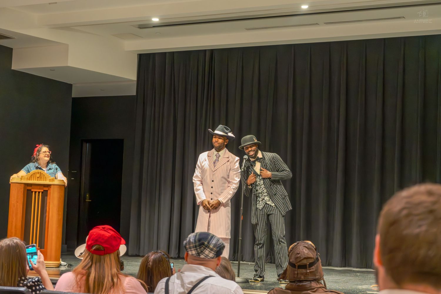 Two performers in vintage suits and hats stand on stage during a theatrical performance.