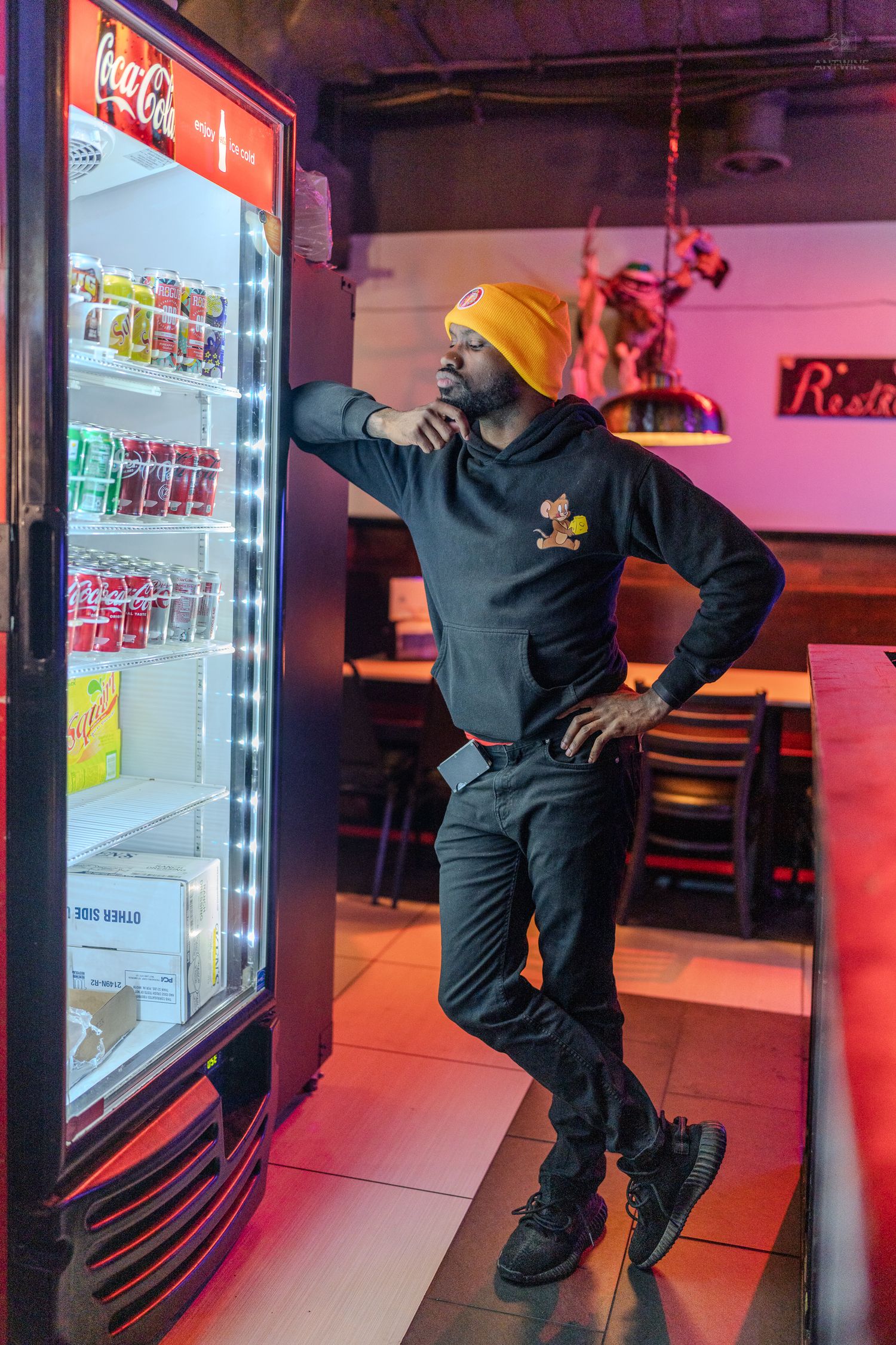 Stylish man in black hoodie and yellow beanie posing beside a glowing Coca-Cola drink cooler in a bar.