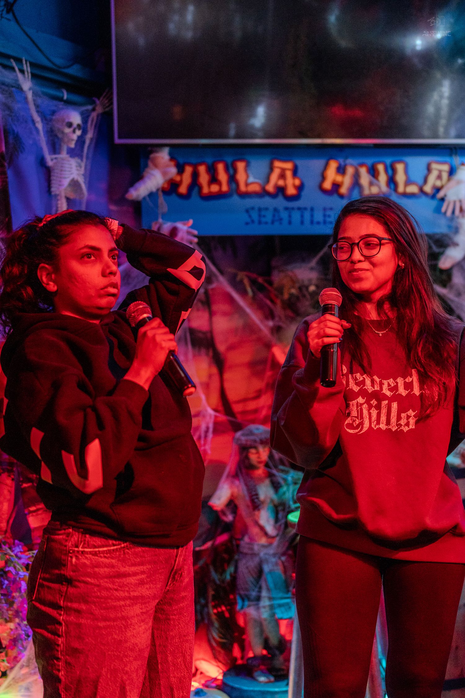 Two women singing karaoke with microphones at Hula Hula Seattle bar under red lighting.