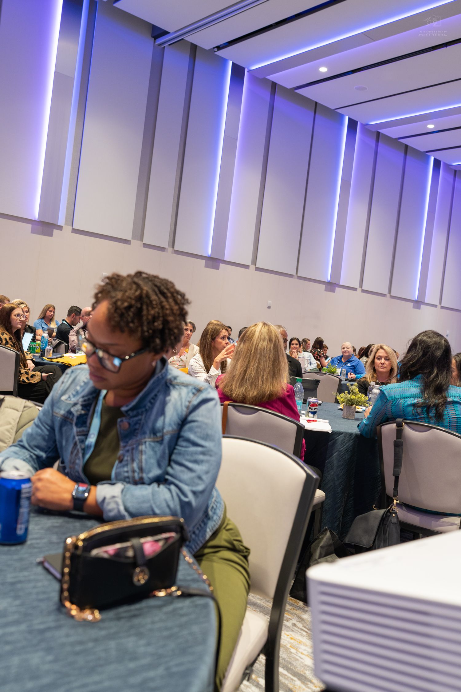 A diverse crowd of attendees gather at a large conference banquet hall with purple lighting.