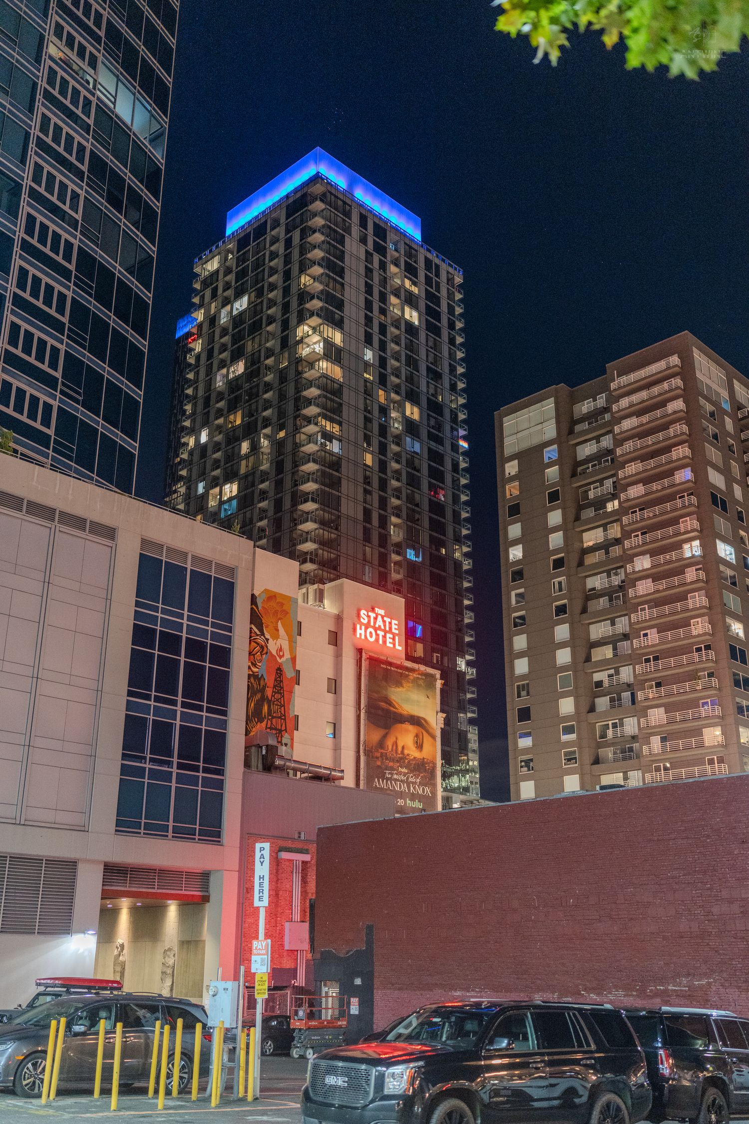 Night cityscape featuring the State Hotel sign, illuminated skyscrapers with blue rooftop lighting, and parked cars in Seattle.