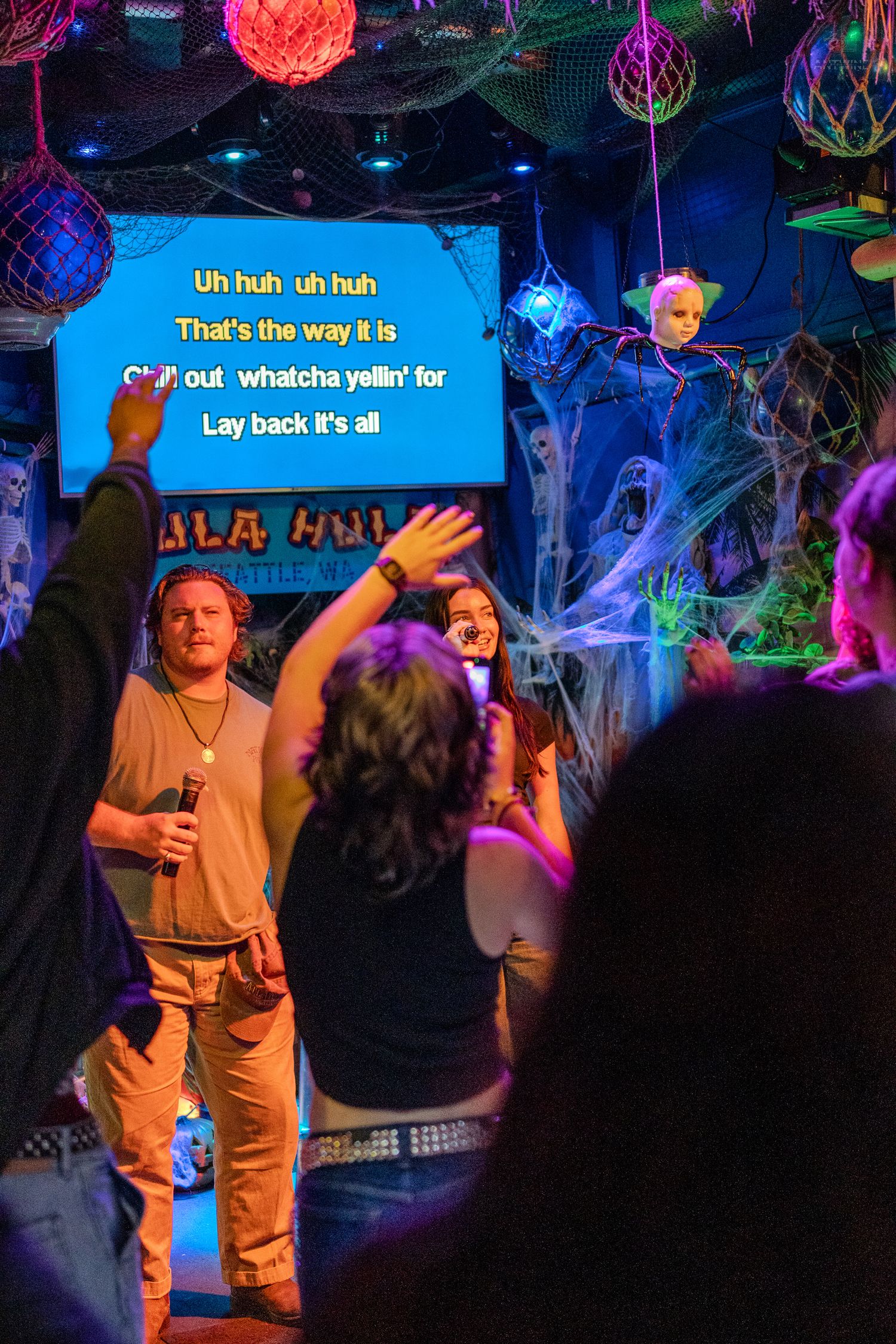 Performers and crowd singing karaoke at Halloween-decorated Hula Hula bar in Seattle with raised hands.