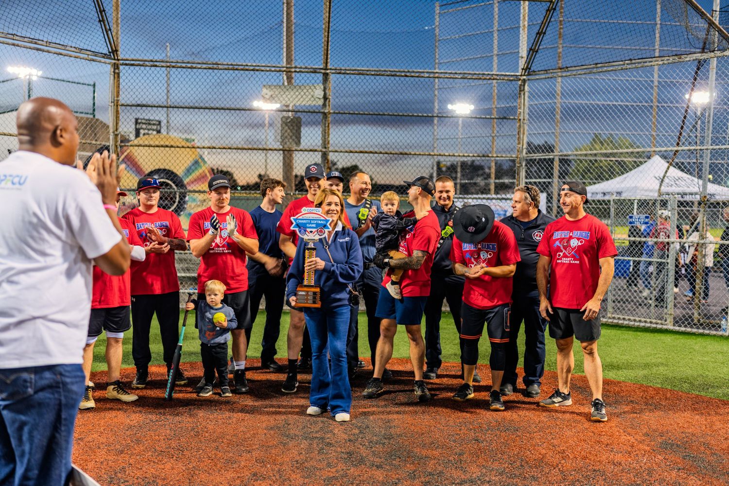 Softball team in red shirts celebrates championship win holding trophy on field at night under stadium lights.