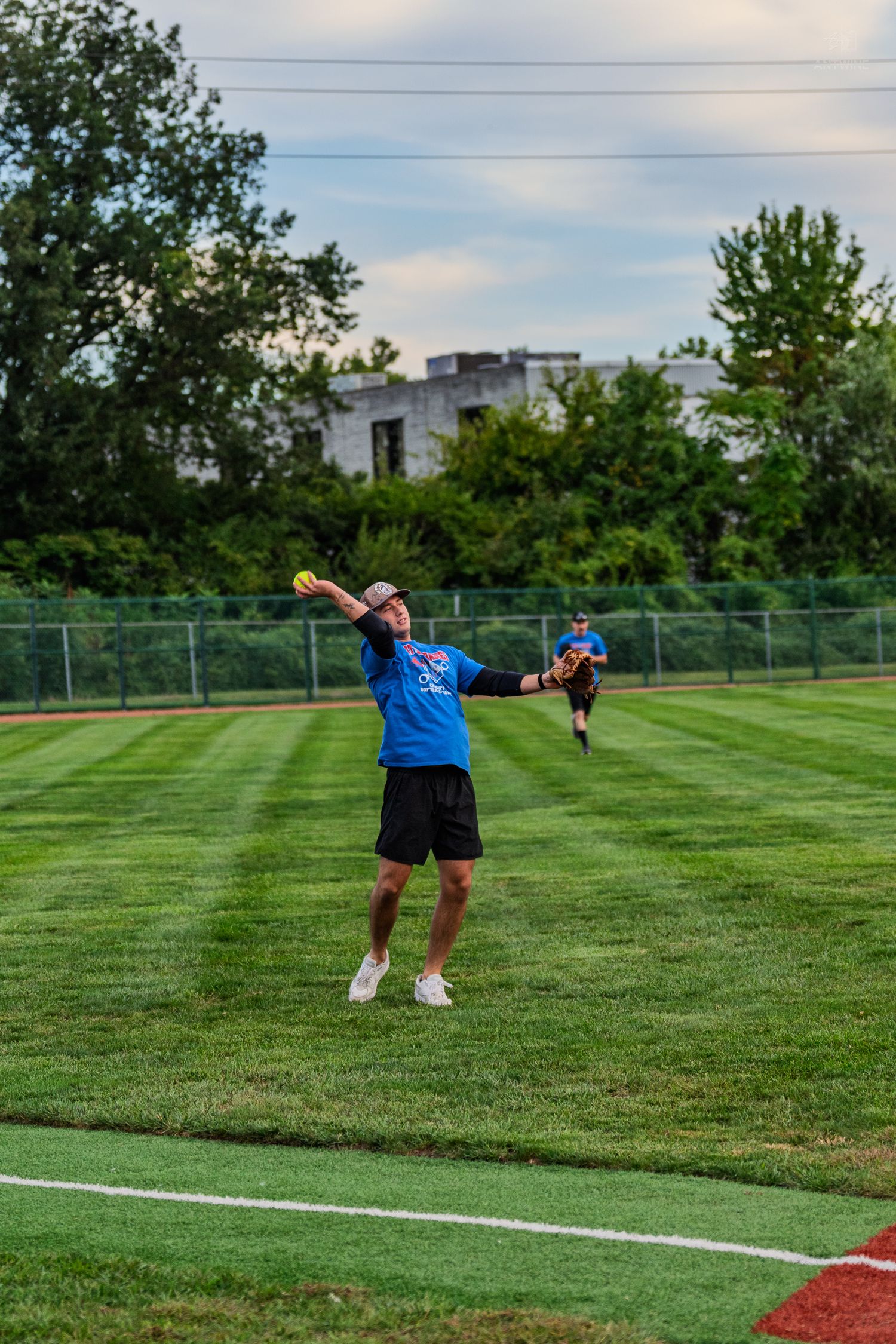 Male softball player in blue jersey winding up to throw yellow ball across green outfield grass.