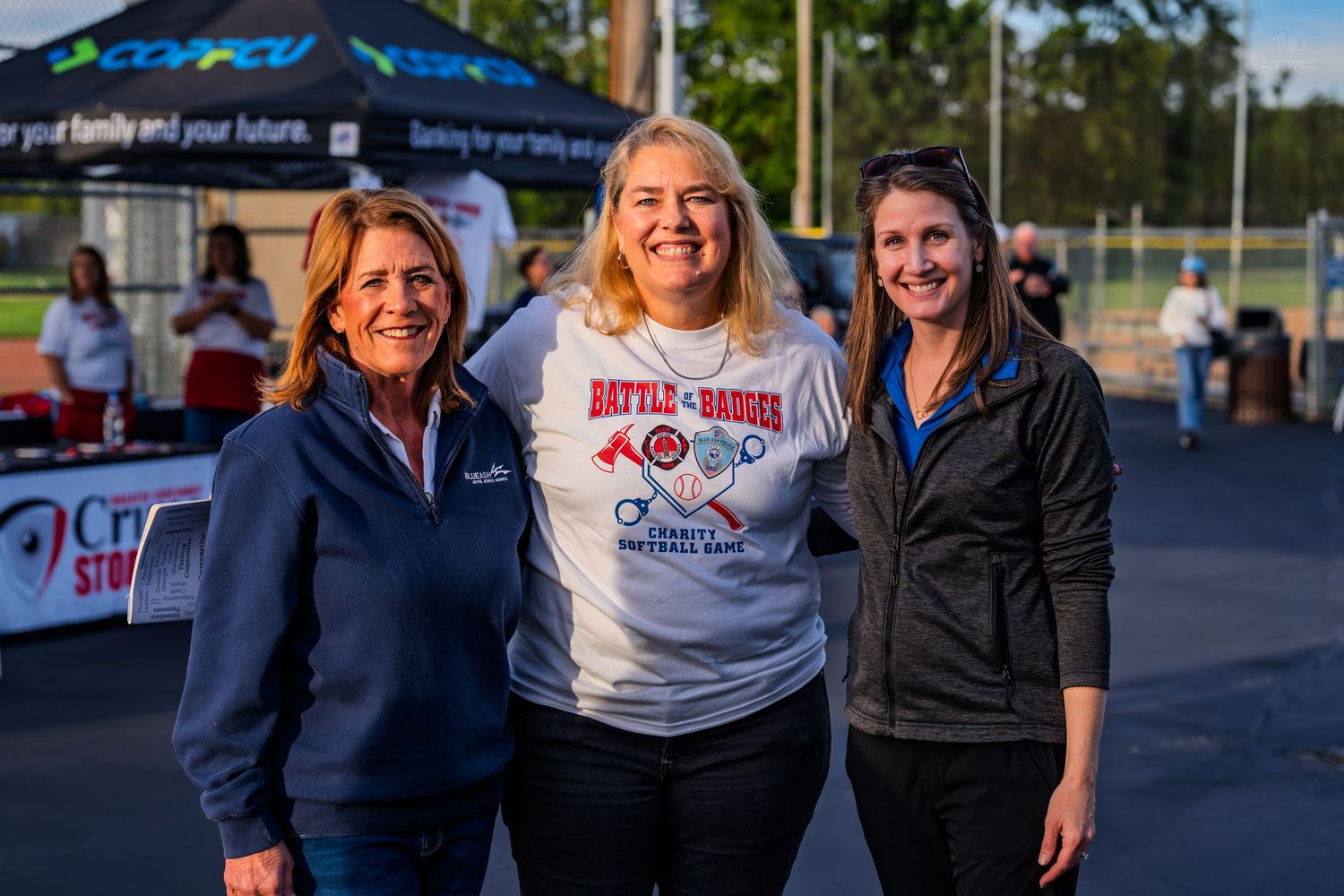 Three smiling women pose together outdoors at a Battle of the Badges charity softball game event.