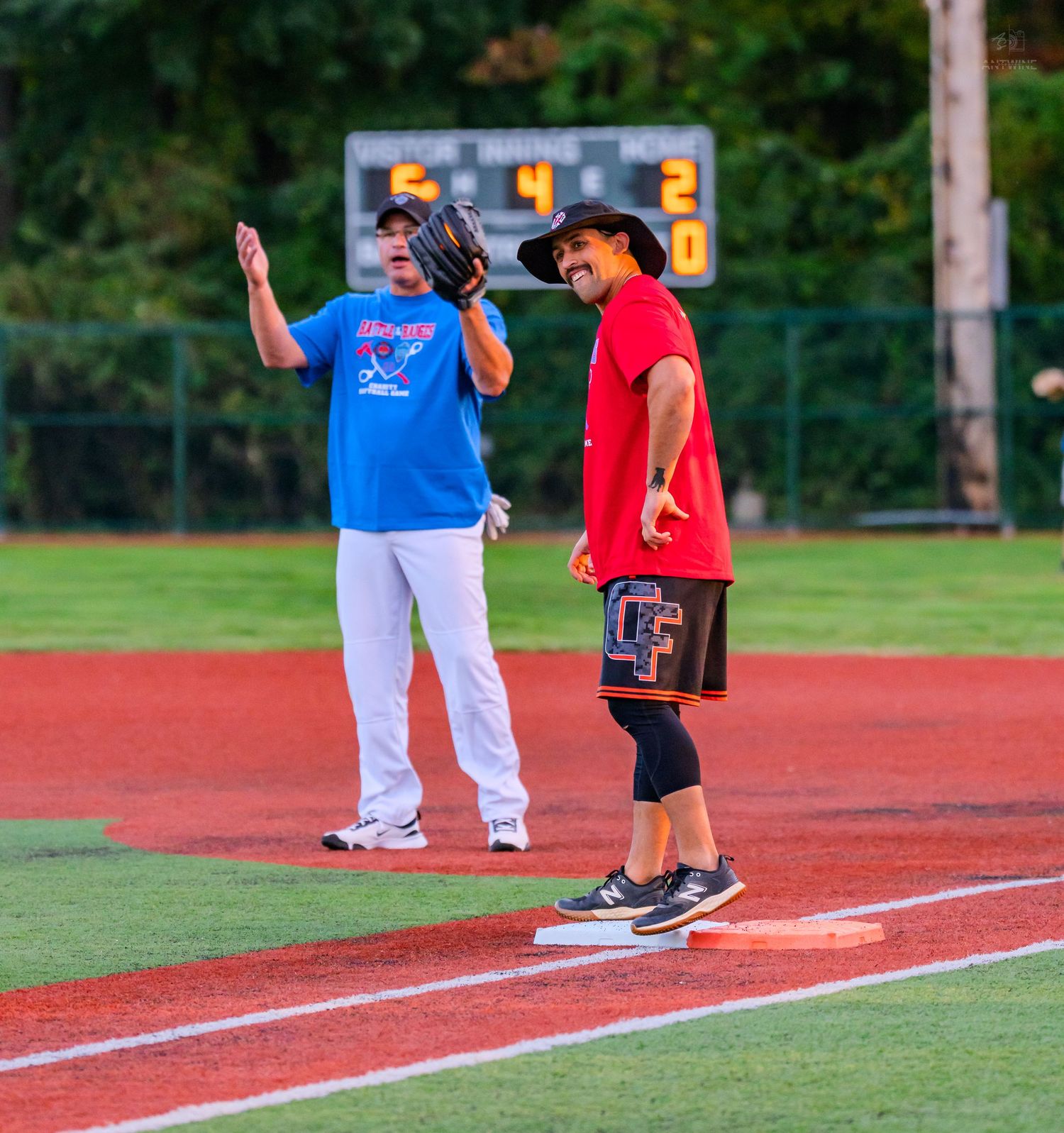 Two players share a laugh at first base during a recreational softball game under stadium lights.