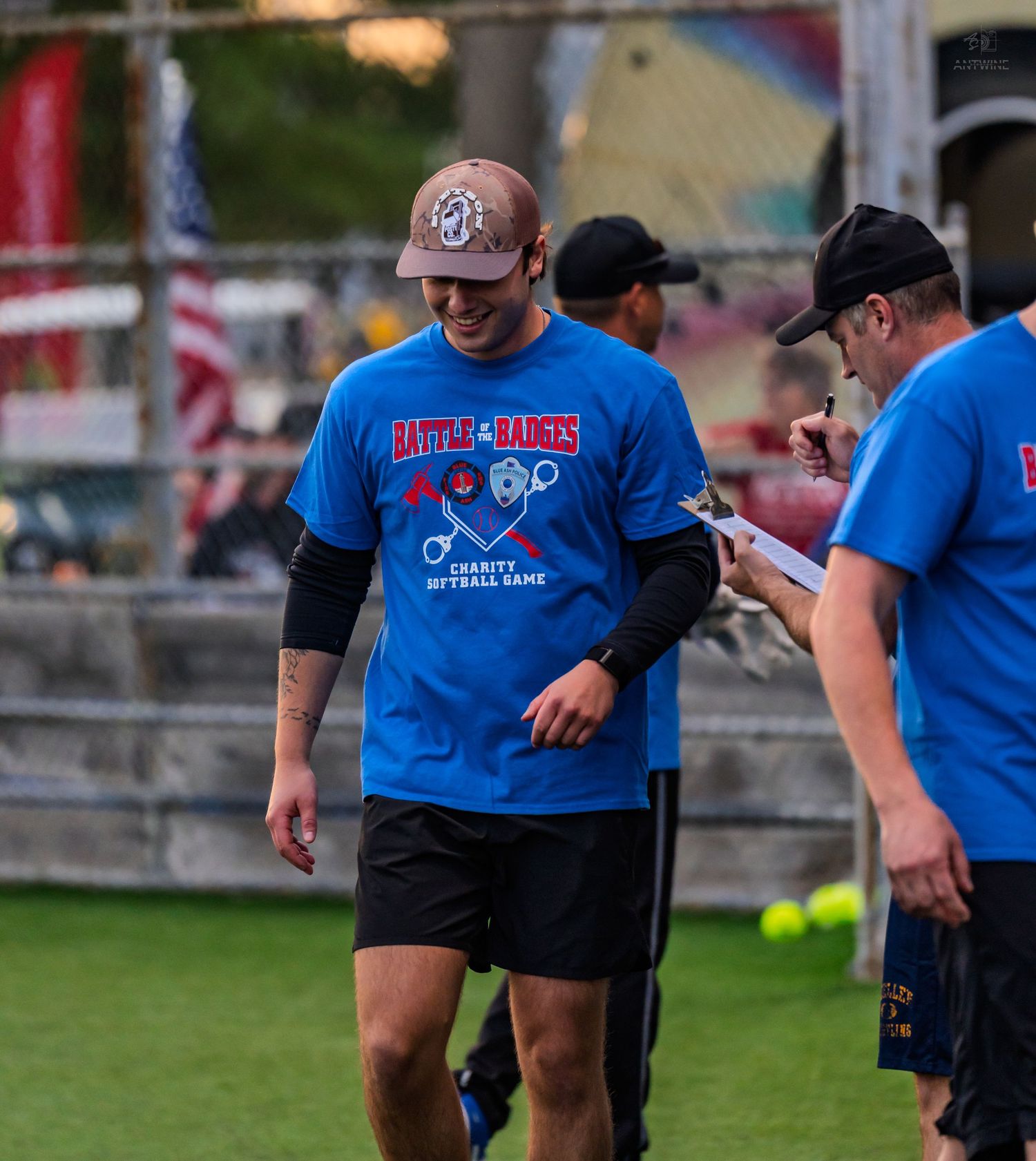A smiling man in a blue charity softball game t-shirt walks on a green field during a fundraiser event.