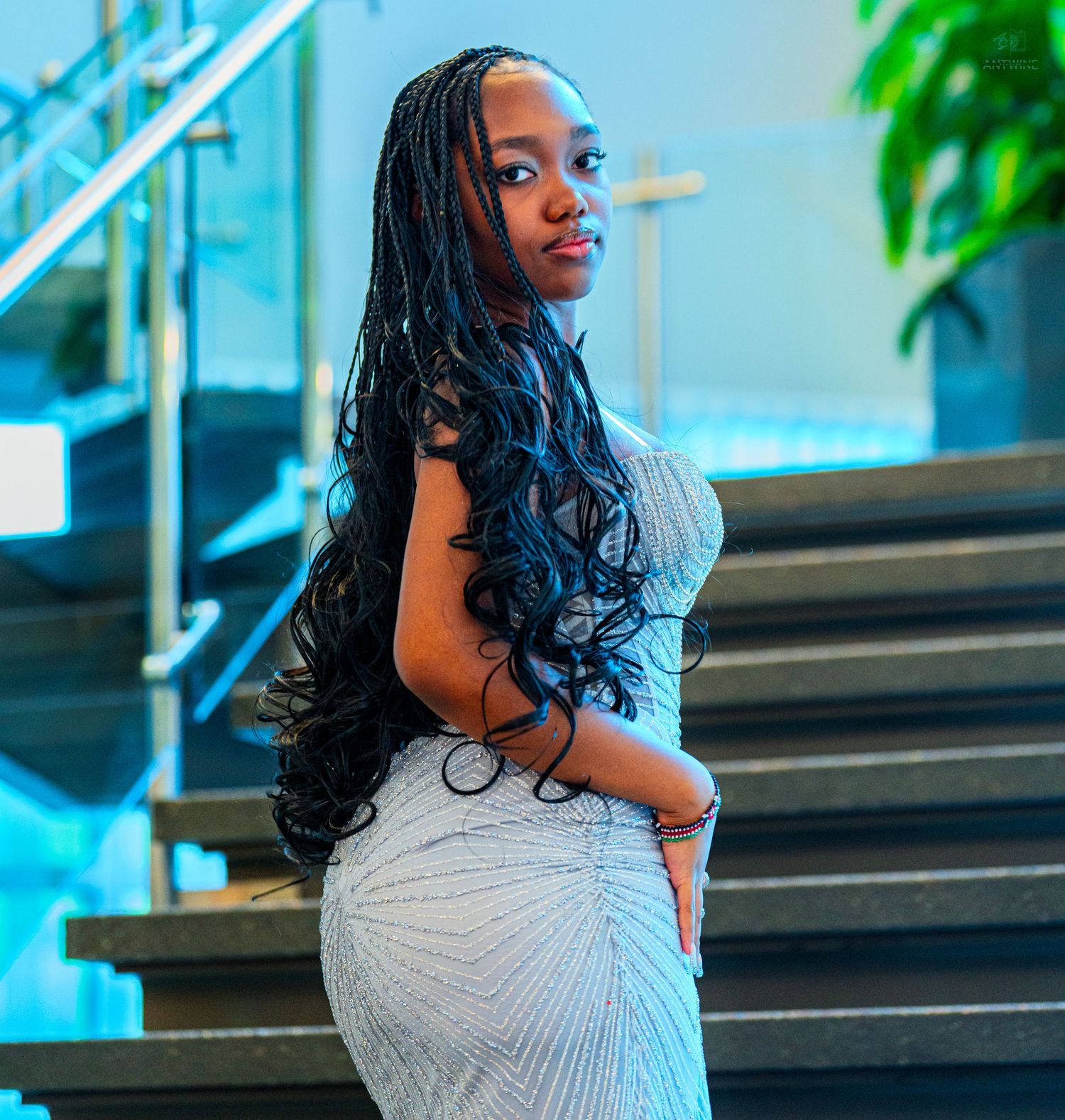 Young woman in silver sequin gown posing elegantly on modern staircase from side.