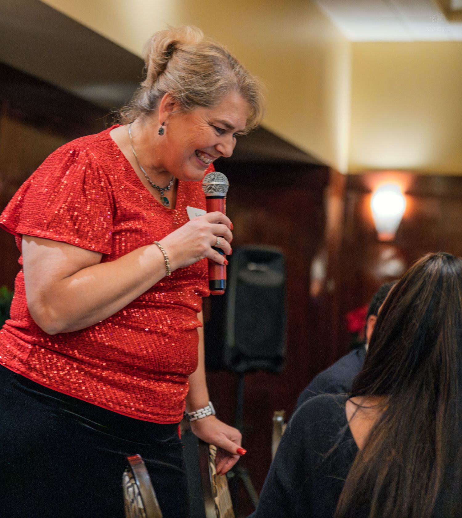 Woman in red sequin top holds microphone, leaning toward seated guest at indoor event.