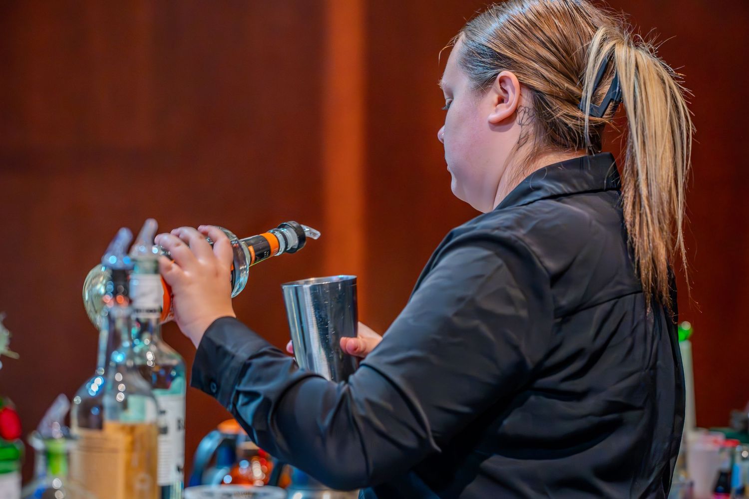 Female bartender in black jacket mixing cocktails with a shaker at a professional bar setup.