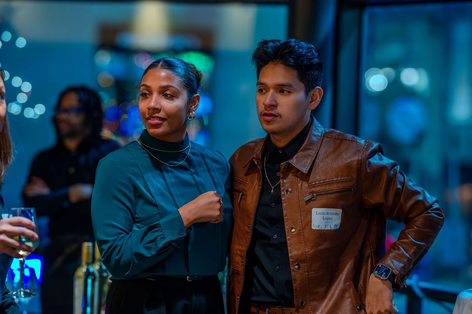 Two young adults posing together at a social event, woman in teal top and man in brown leather jacket.