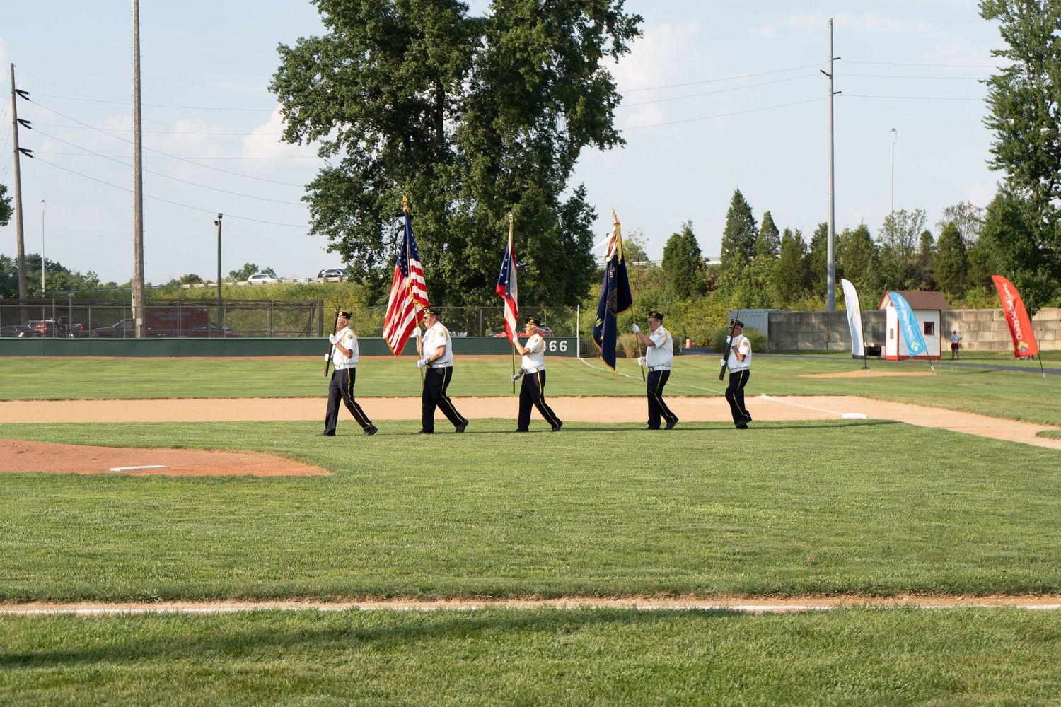 Color guard marching with American flag on baseball field during outdoor ceremony on a sunny day.