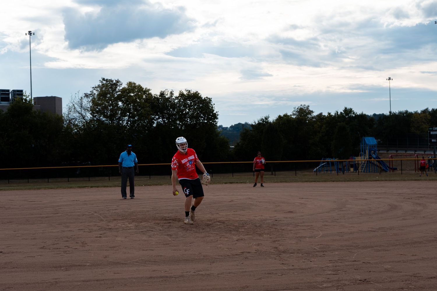 Players on a sandy softball field during an evening game with cloudy skies and trees in the background.
