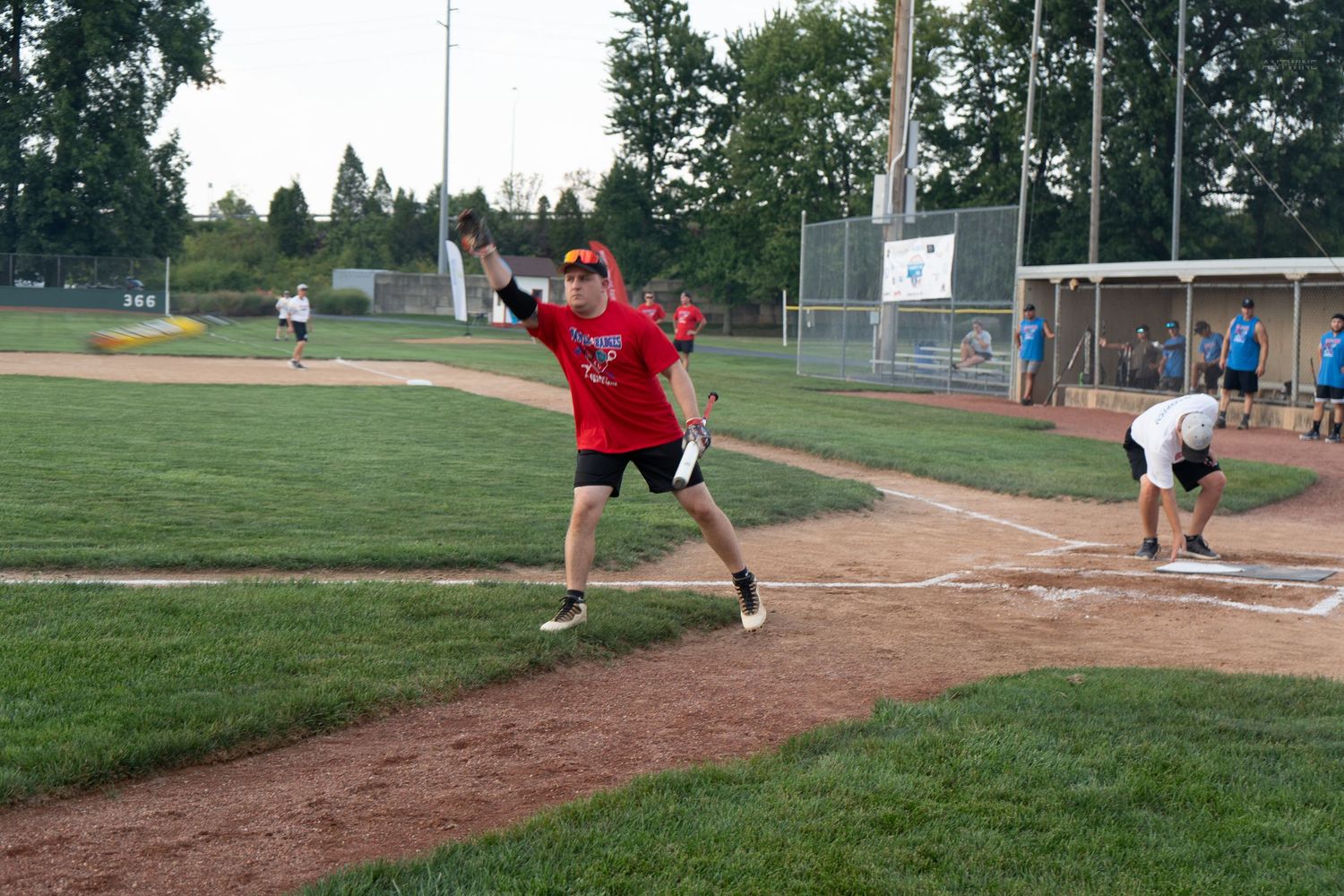 Baseball player in red uniform throwing ball on dirt infield during an outdoor recreational baseball game at dusk.