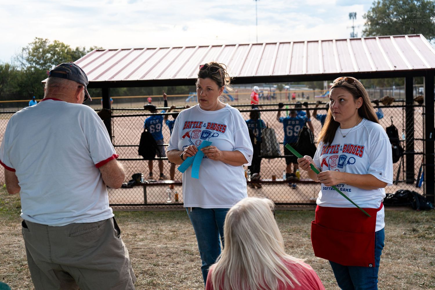 Group of people in matching white t-shirts socializing at an outdoor softball field event during daytime.