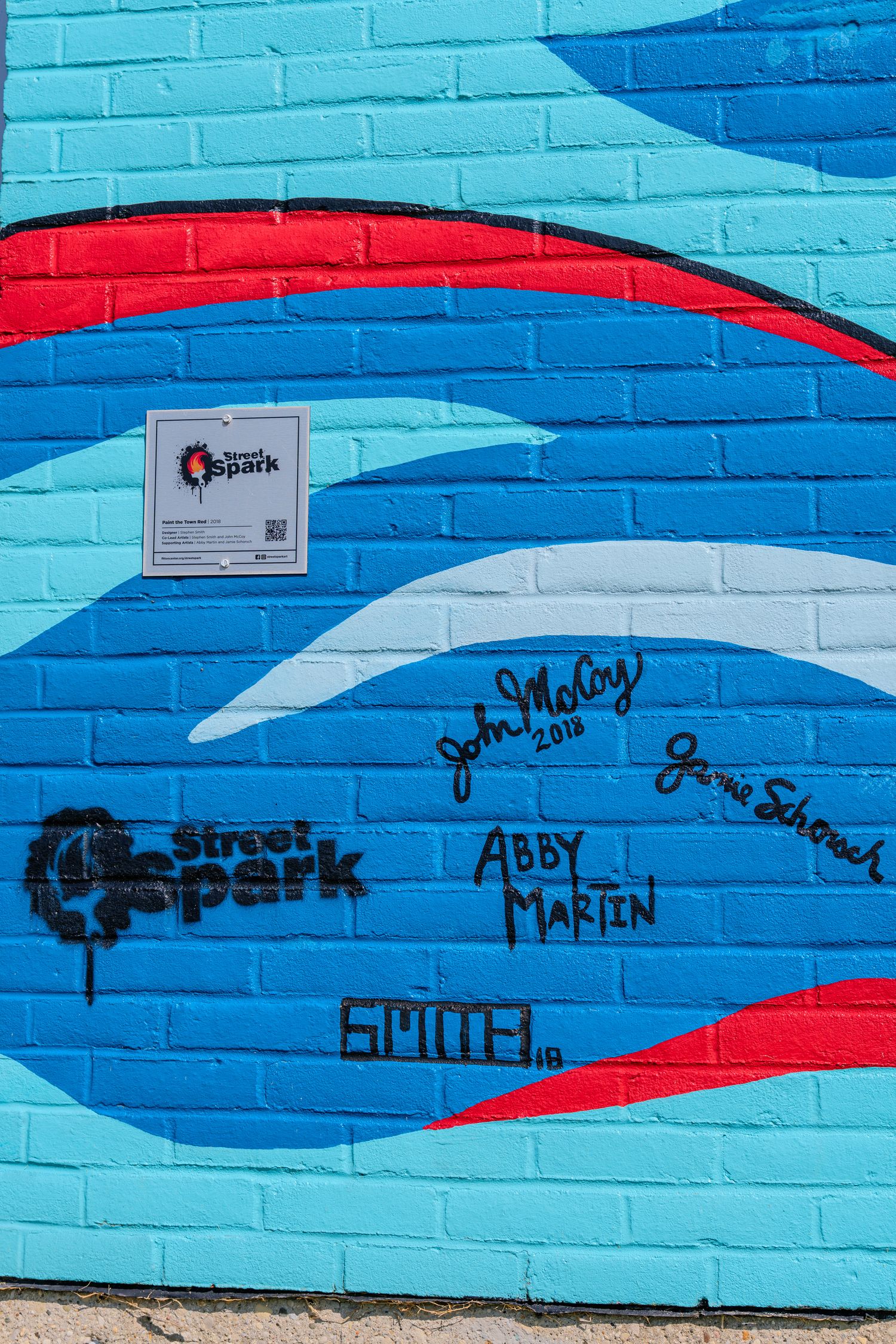 Colorful blue brick wall mural with red accents featuring Street Spark graffiti art and signatures including Abby Martin.