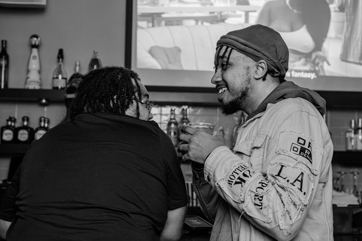 Two men laughing and toasting drinks at a bar in a black and white photo.
