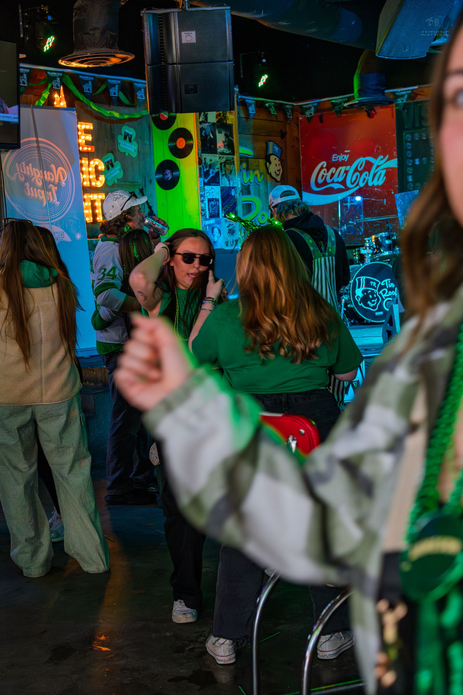 People in green outfits celebrate St. Patrick's Day at a bar with Coca-Cola signs and neon lights.