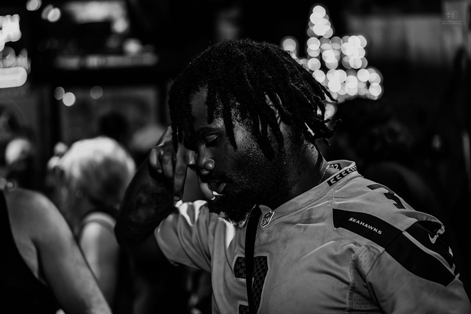 Black and white photo of a young man with dreadlocks wearing a jersey in a crowded nighttime outdoor setting.