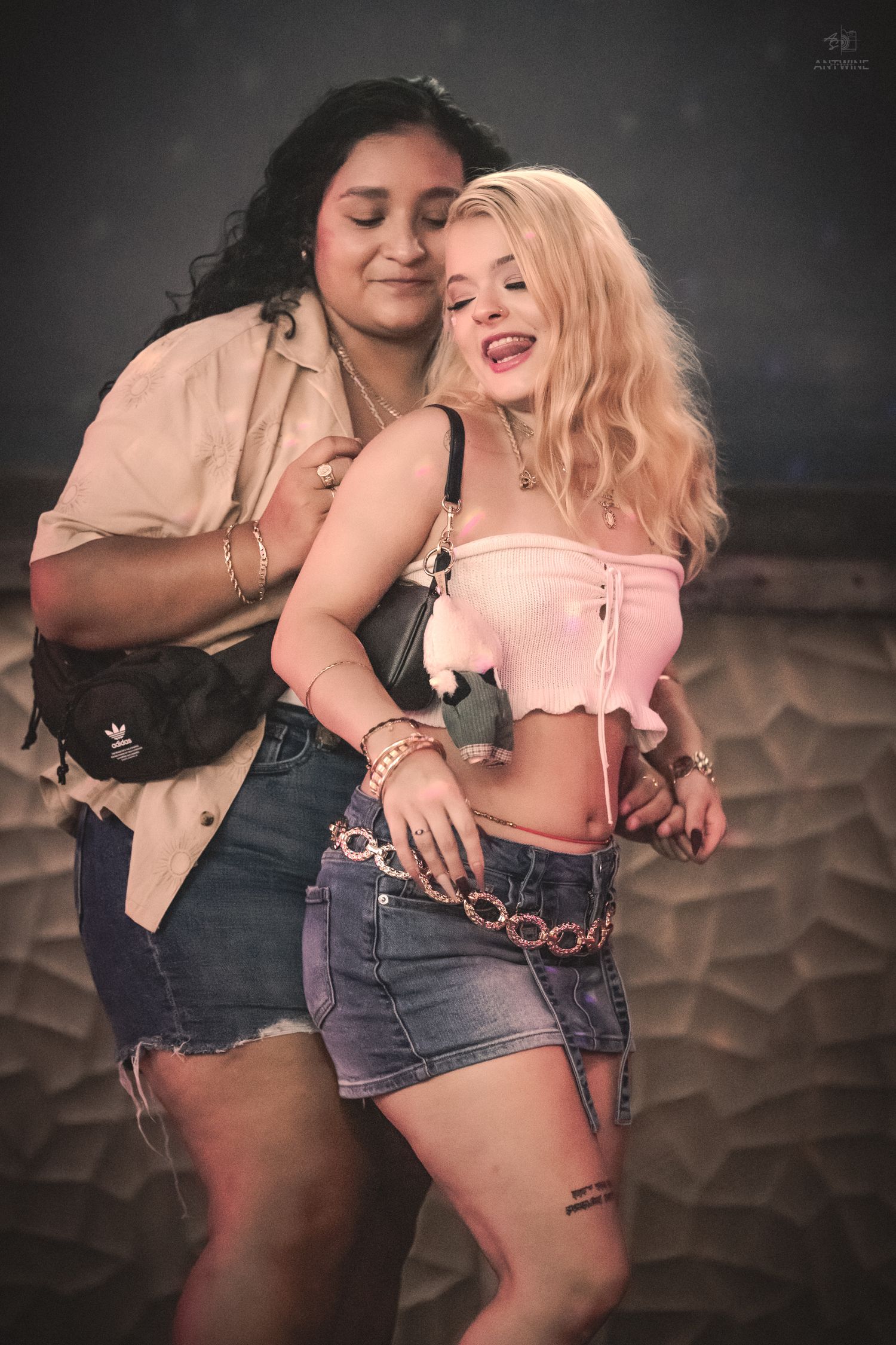 Two young women posing together at a party, one in a beige shirt with fanny pack, other in pink crop top.