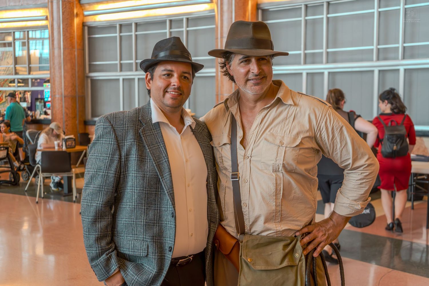 Two men in vintage-style costumes pose together indoors, one in a suit and fedora, another in explorer attire.