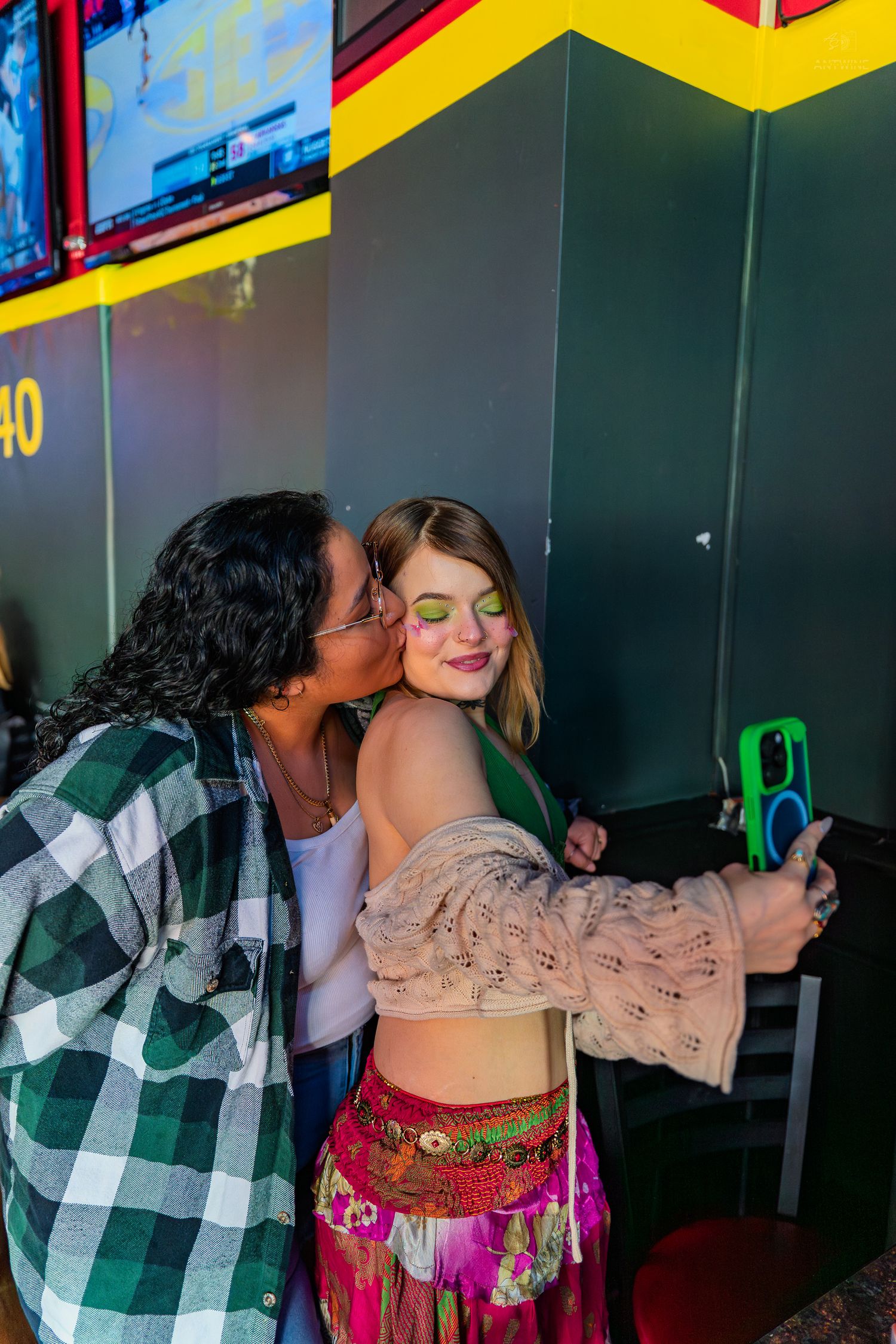 Two women take a selfie, one kissing the other's cheek at a colorful arcade bar.