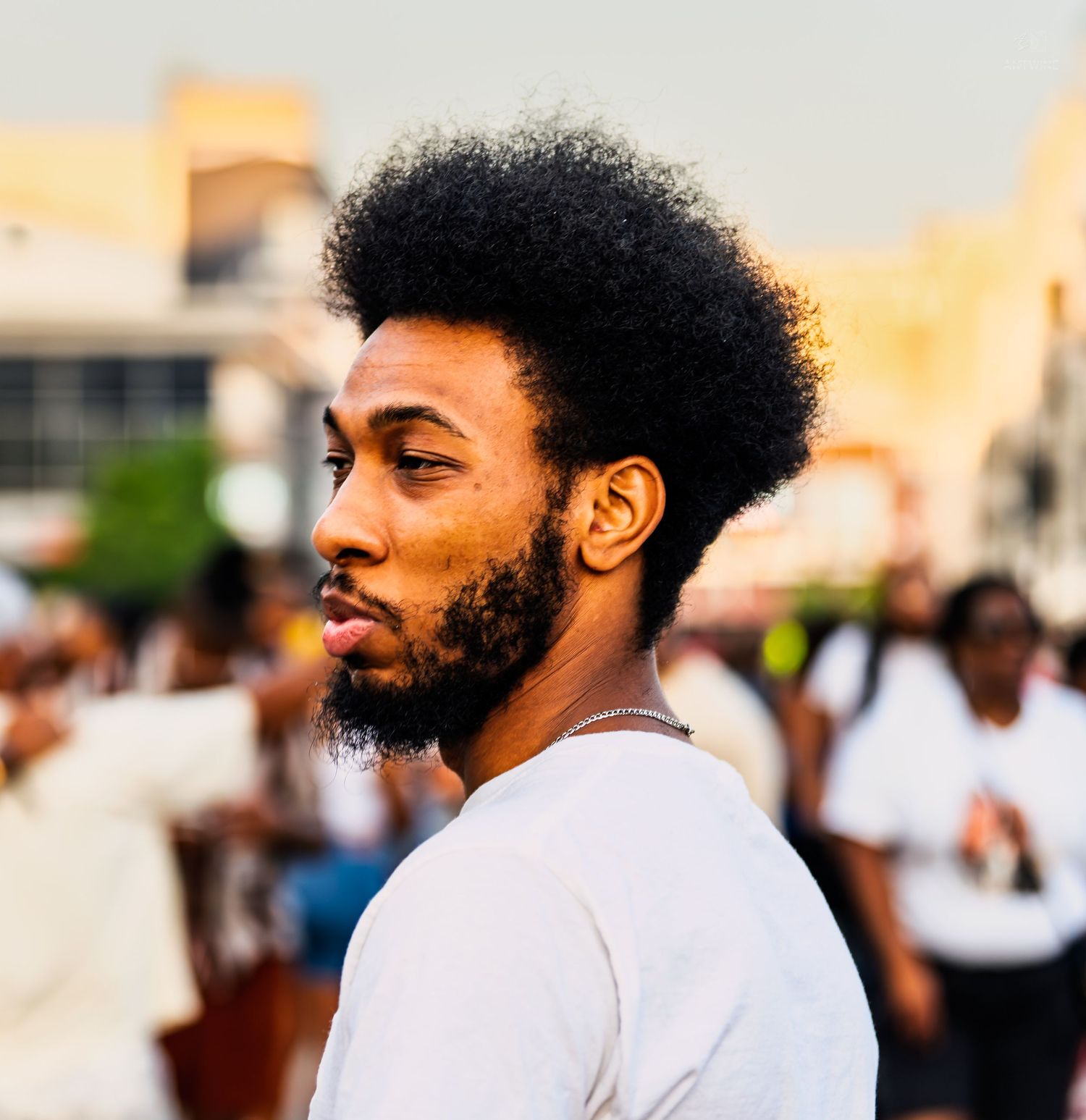 Young Black man with afro and beard in white shirt looks over shoulder at outdoor urban gathering in golden hour light.