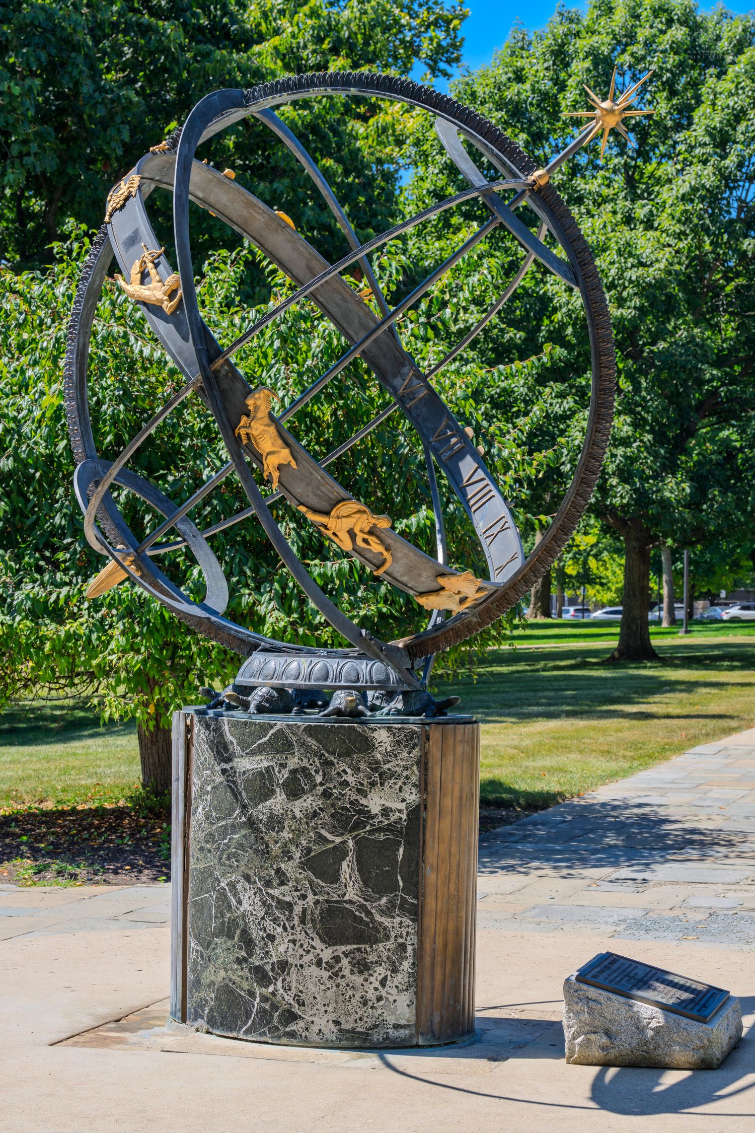 Ornate armillary sphere sundial sculpture on marble pedestal in a lush green park setting.